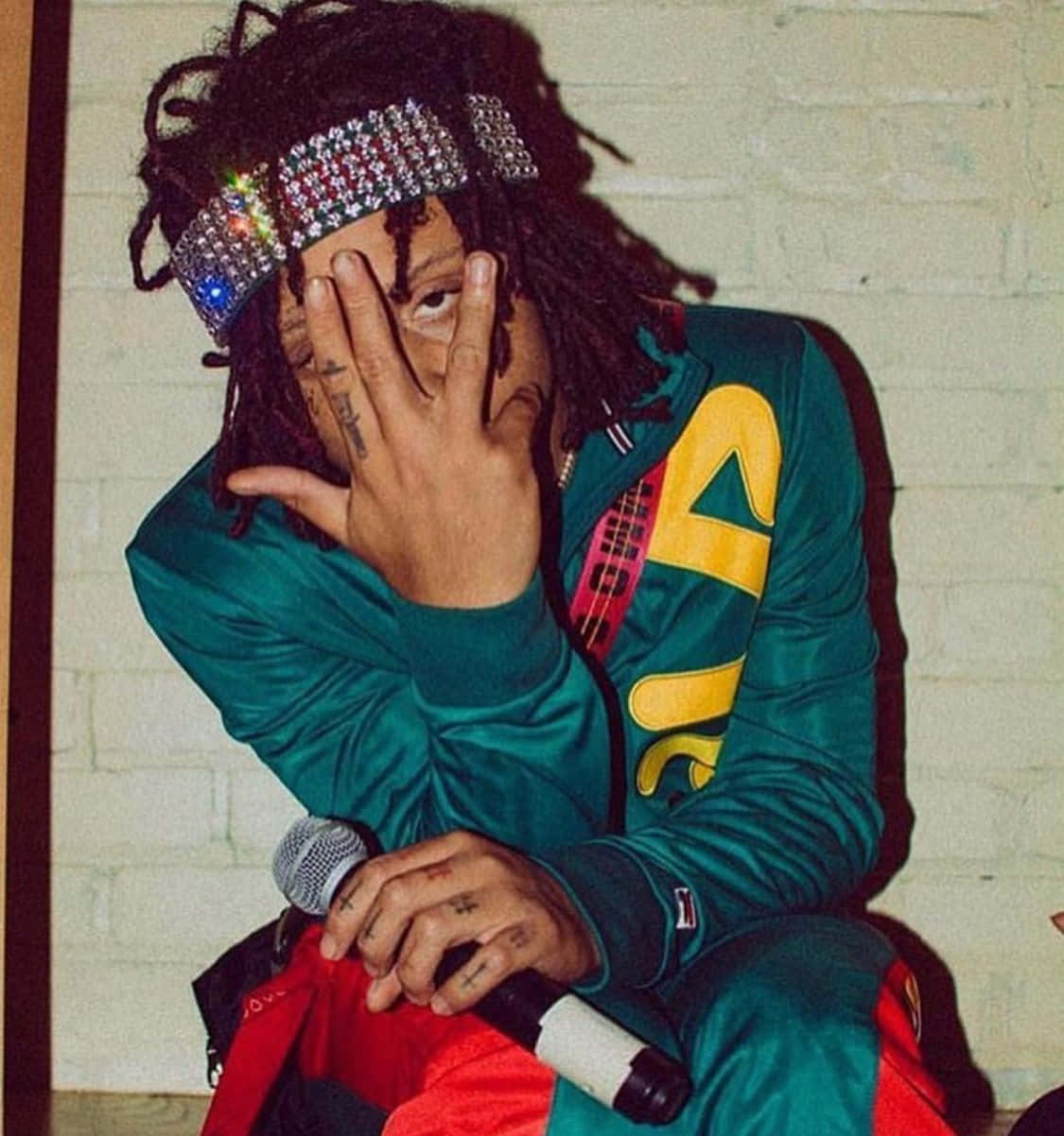 trippie redd 桌布 壁紙 — 一個留著辮子、戴著珠寶頭帶的人，部分遮住臉，拿著麥克風靠著磚牆。