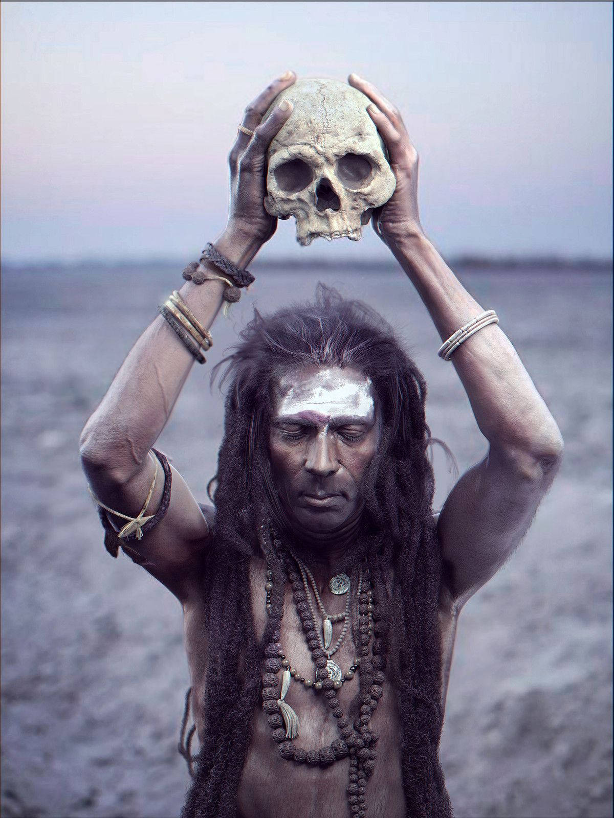 Aghori 桌布 壁紙 — 一個留著辮子、臉上塗著顏料的男子，在暗淡的天空下將一個人類頭骨高舉過頭頂。