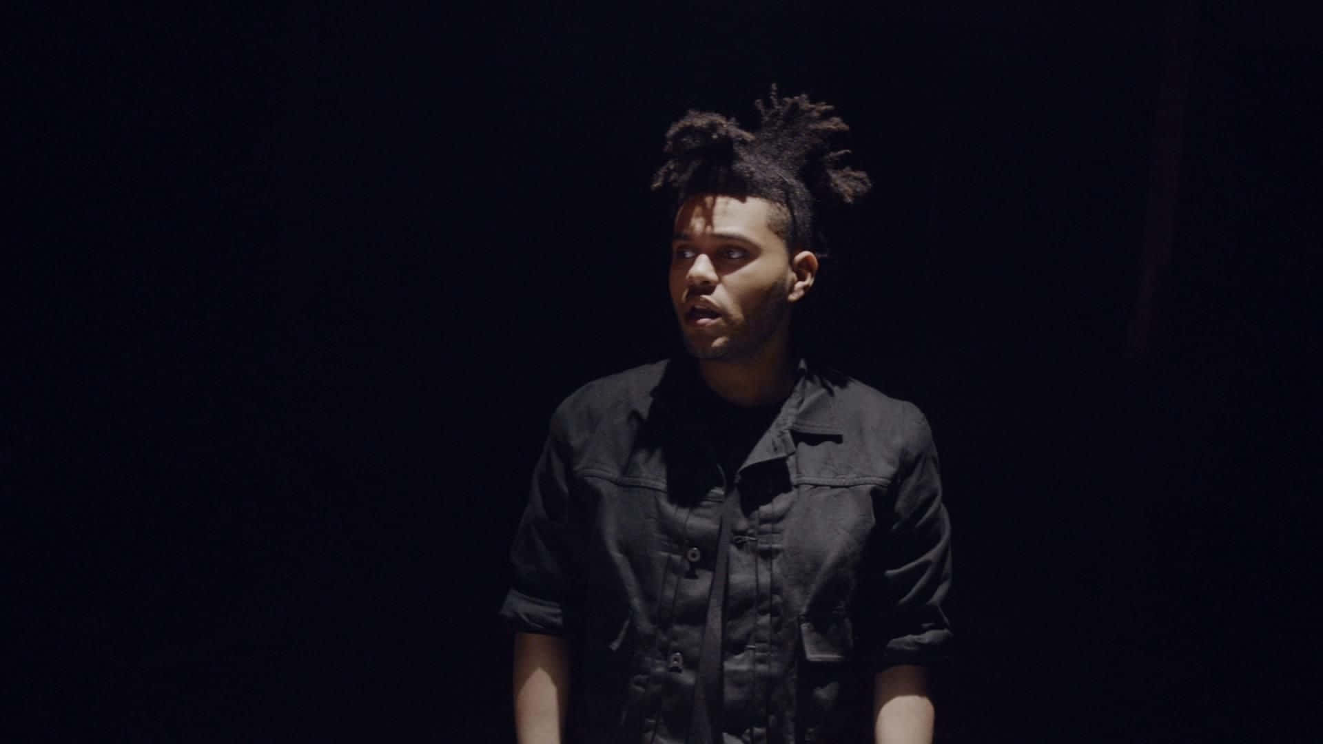 The Weeknd 桌布 壁紙 — 一個留著辮子頭的男人站在黑暗中，被單一聚光燈照亮，穿著黑色襯衫。