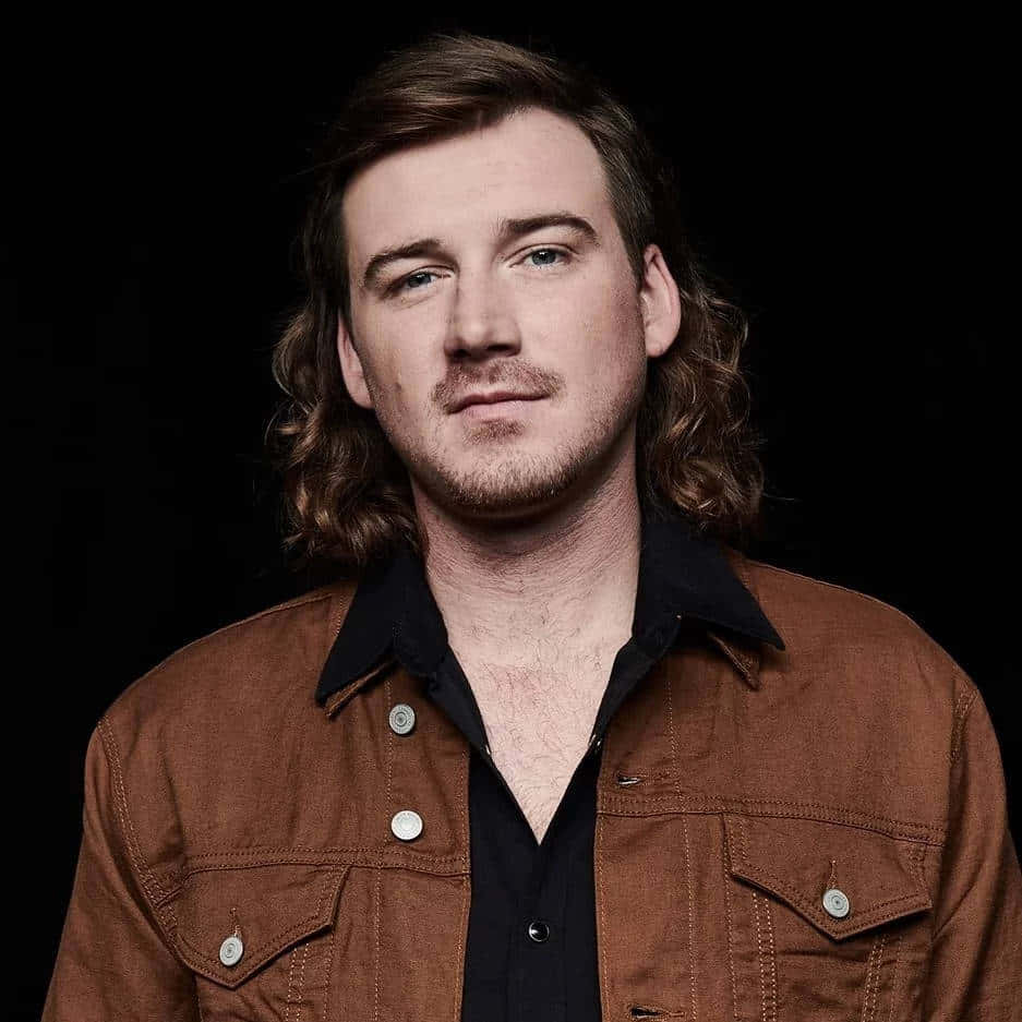 Morgan Wallen 桌布 壁紙 — 一個留著長波浪棕色頭髮和鬍鬚的男人，在黑色背景前，穿著棕色牛仔外套內搭黑色襯衫。