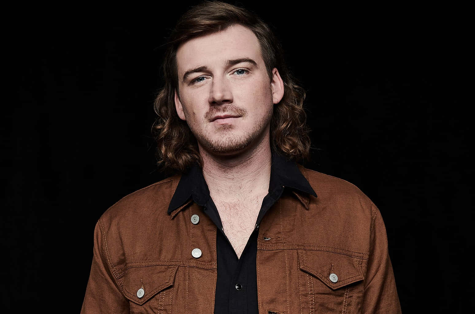 Morgan Wallen 桌布 壁紙 — 一個留著長波浪棕色頭髮和鬍鬚的男人，在黑色背景前穿著黑色襯衫外搭棕色外套。