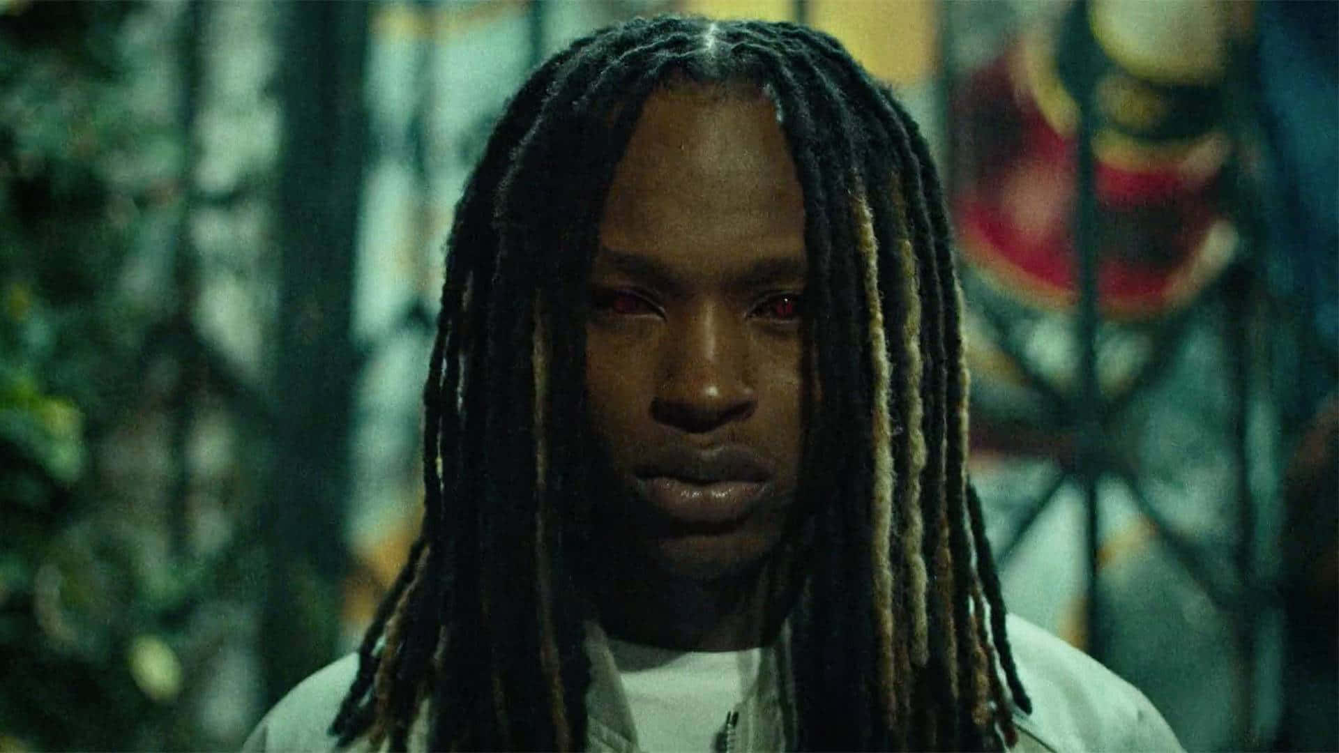 King Von 桌布 壁紙 — 一個留著長辮子的男人在昏暗模糊的城市環境中，背景有塗鴉，專注地向前凝視。