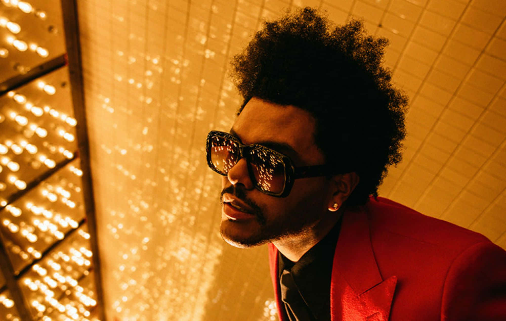 The Weeknd After Hours 桌布 壁紙 — 一個留著非洲式髮型、戴著太陽眼鏡的男人，身穿紅色西裝外套，站在閃閃發光的金色瓷磚牆前。