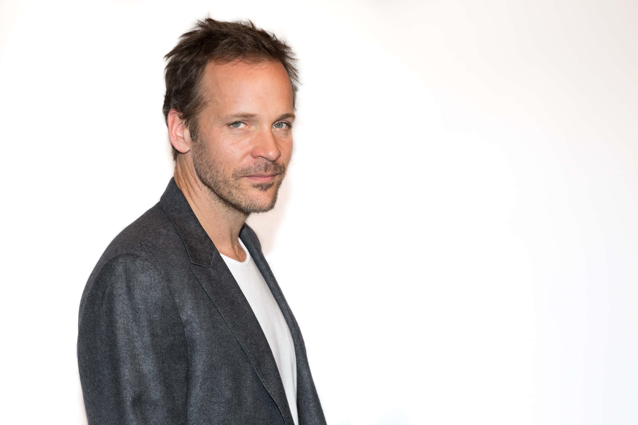 Peter Sarsgaard 桌布 壁紙 — 一個留著鬍渣、頭髮凌亂的男人，在純白背景前穿著深色西裝外套，內搭白色T恤。