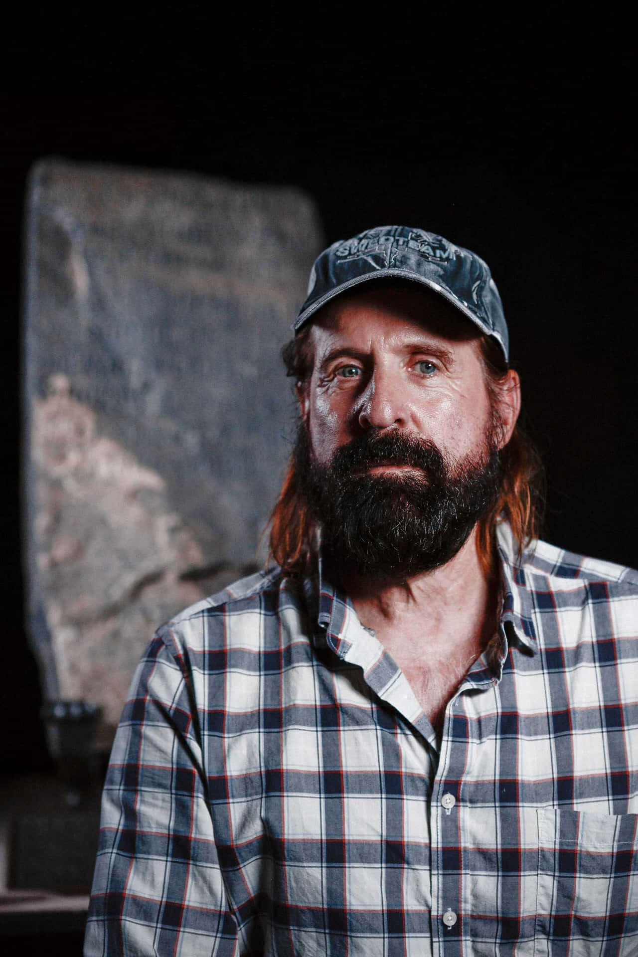 Peter Stormare 桌布 壁紙 — 一個留長鬍鬚的男人，長髮，穿著格仔襯衫和帽子，背景黑暗，身後有一塊石板。