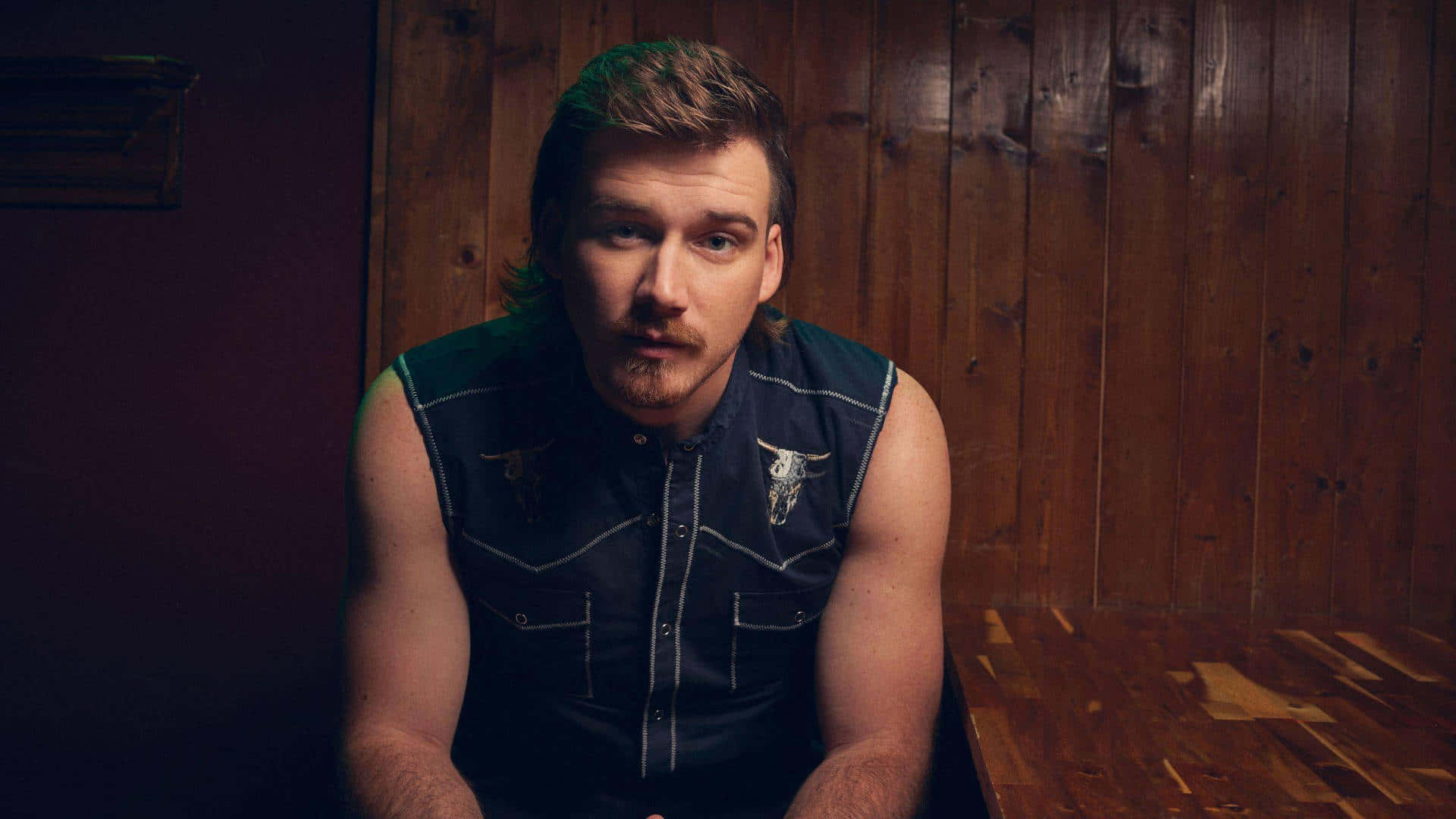 Morgan Wallen 桌布 壁紙 — 一個留鬍子、頭髮帶綠色調的男人坐在昏暗房間的木牆旁。