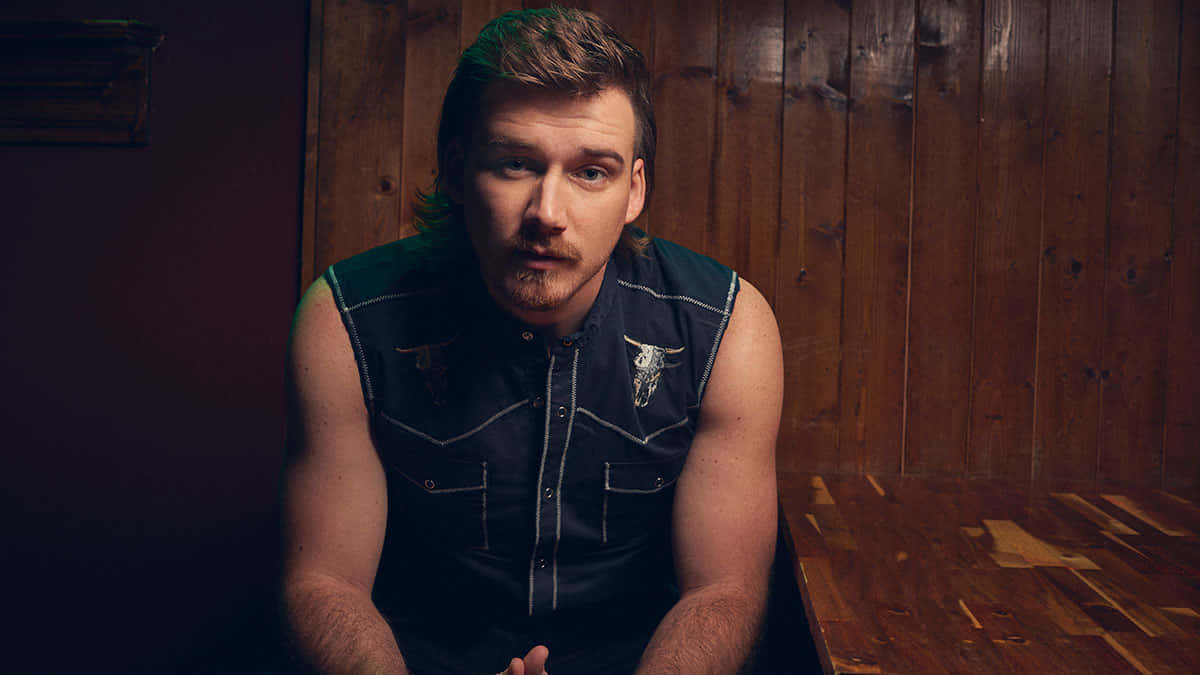 Morgan Wallen 桌布 壁紙 — 一個留鬍子、頭髮帶綠色調的男人坐在昏暗的木房間裡，穿著一件帶有公牛刺繡的深色無袖襯衫。