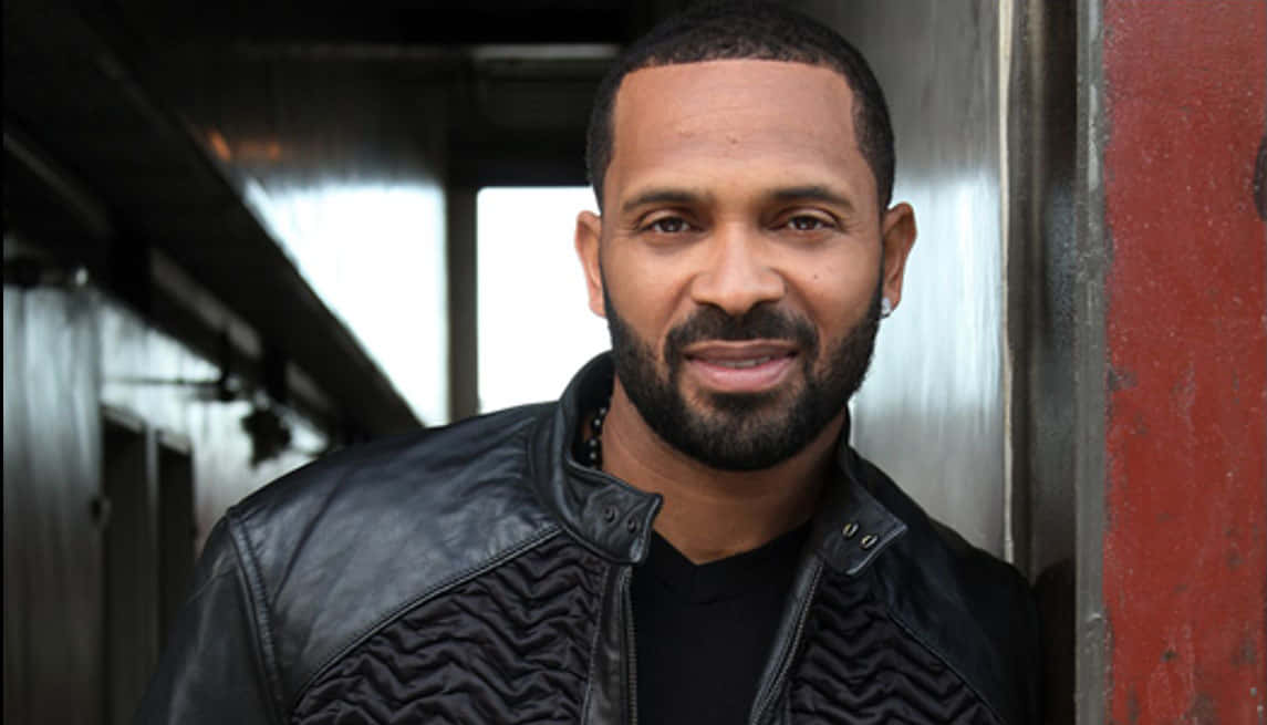 Mike Epps 桌布 壁紙 — 一個留鬍子戴耳環的男人在昏暗的城市走廊裡靠著紅牆微笑。