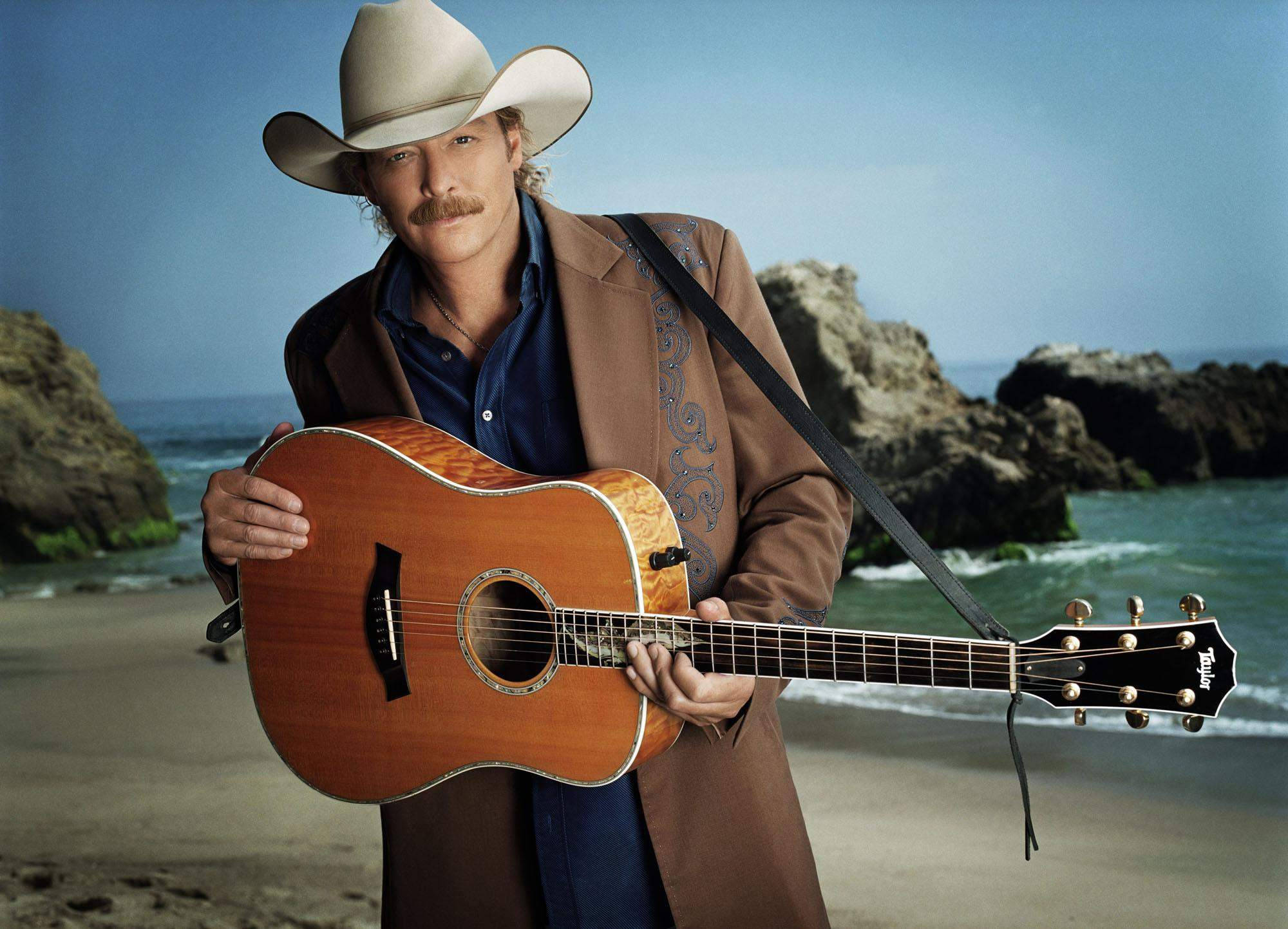 Alan Jackson 桌布 壁紙 — 一個留鬍子的牛仔在岩石海灘上抱著一把原聲結他，身後海浪拍打。
