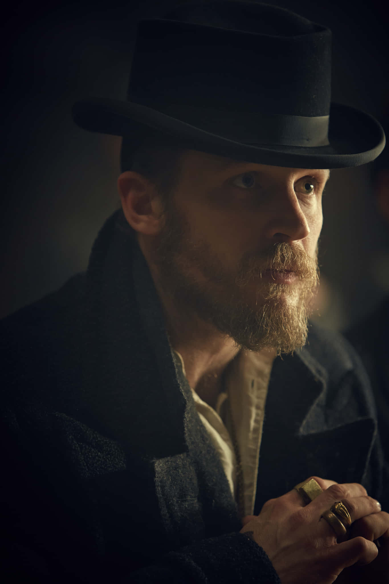 Peaky Blinders 桌布 壁紙 — 一個留鬍子的男人，戴著黑色軟呢帽和外套，在昏暗、情緒化的燈光下，手指上戴著金戒指，專注地凝視著。