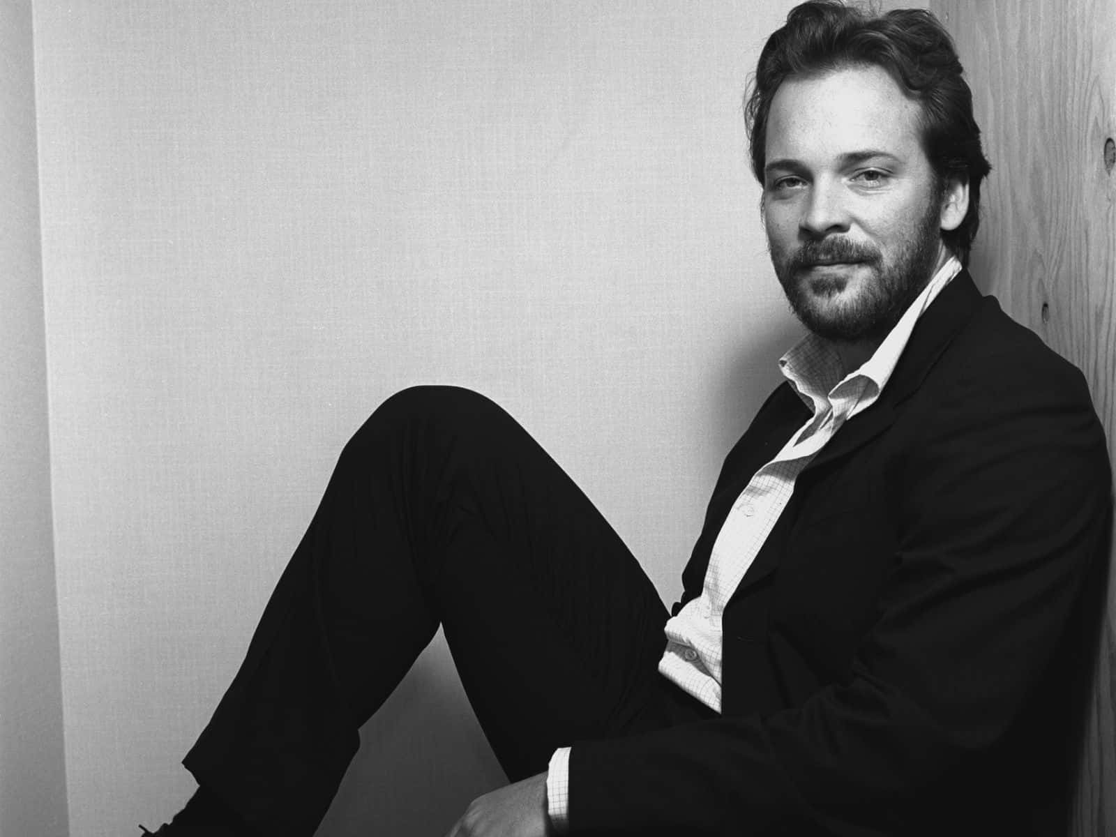Peter Sarsgaard 桌布 壁紙 — 一個留鬍子的男人隨意地靠著紋理牆壁坐著的黑白肖像。
