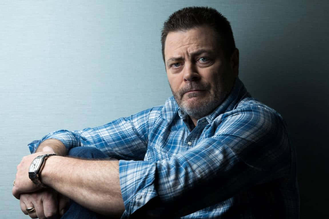 nick offerman 桌布 壁紙 — 一個留鬍鬚、眼神銳利的男人，身穿藍色格仔恤衫，向前傾身坐在柔和的灰色背景前。