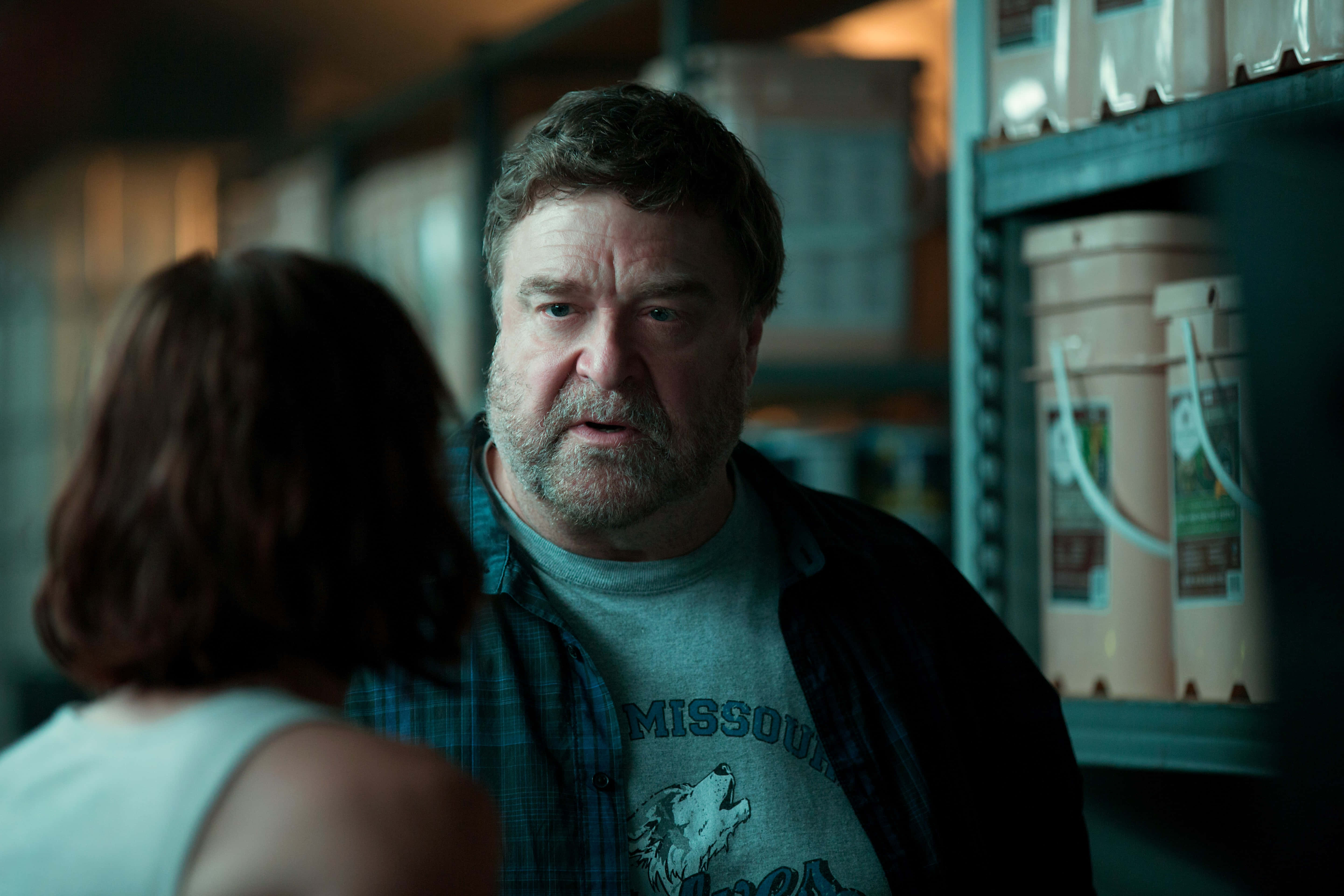 John Goodman 桌布 壁紙 — 一個留鬍鬚、穿著格仔恤衫嘅男人喺燈光昏暗嘅倉庫通道入面專注咁望住一個女人。