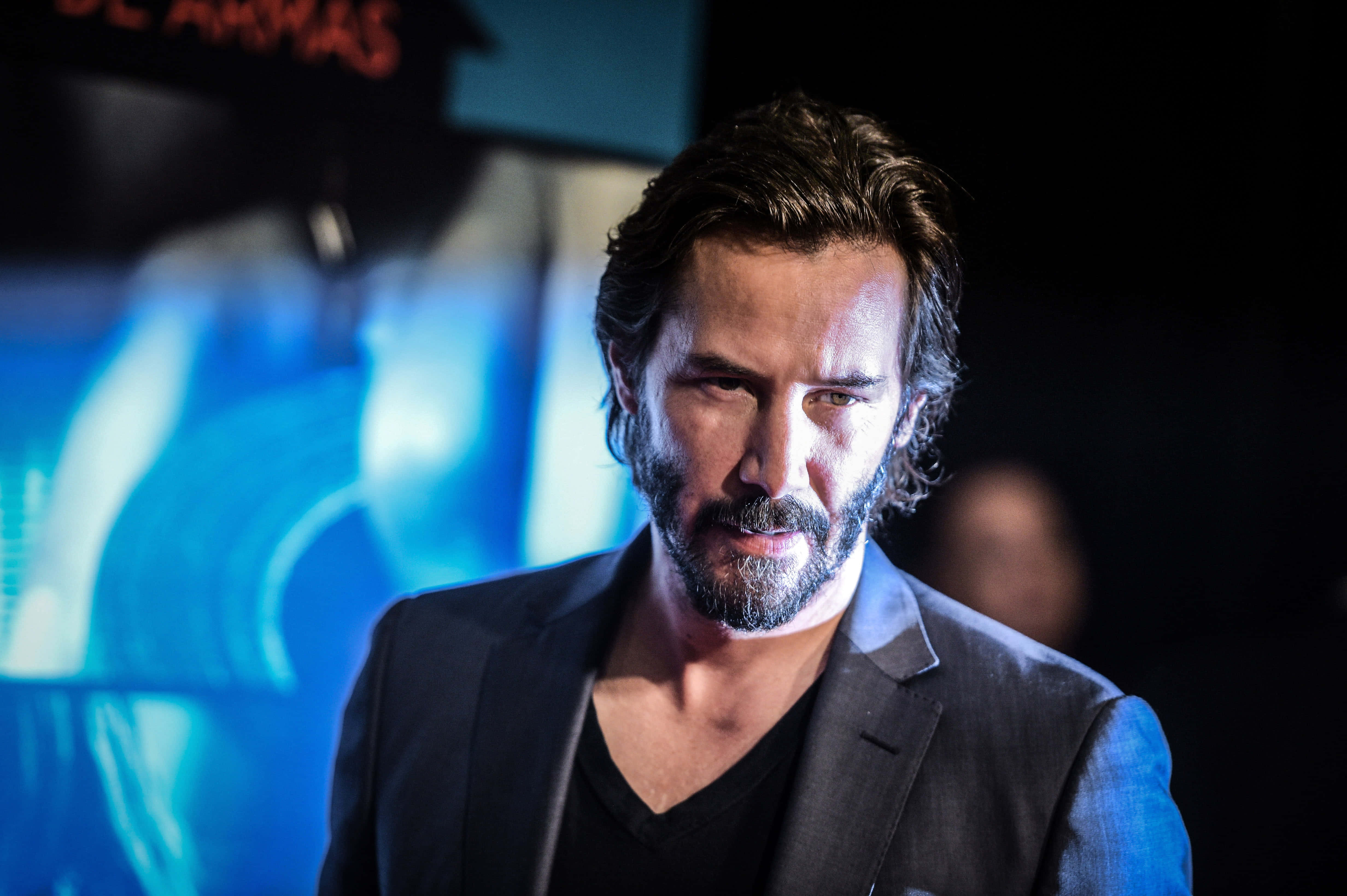 Keanu Reeves 桌布 壁紙 — 一個留鬍鬚、身穿深色西裝的男人，在模糊的藍色背景下凝視著。