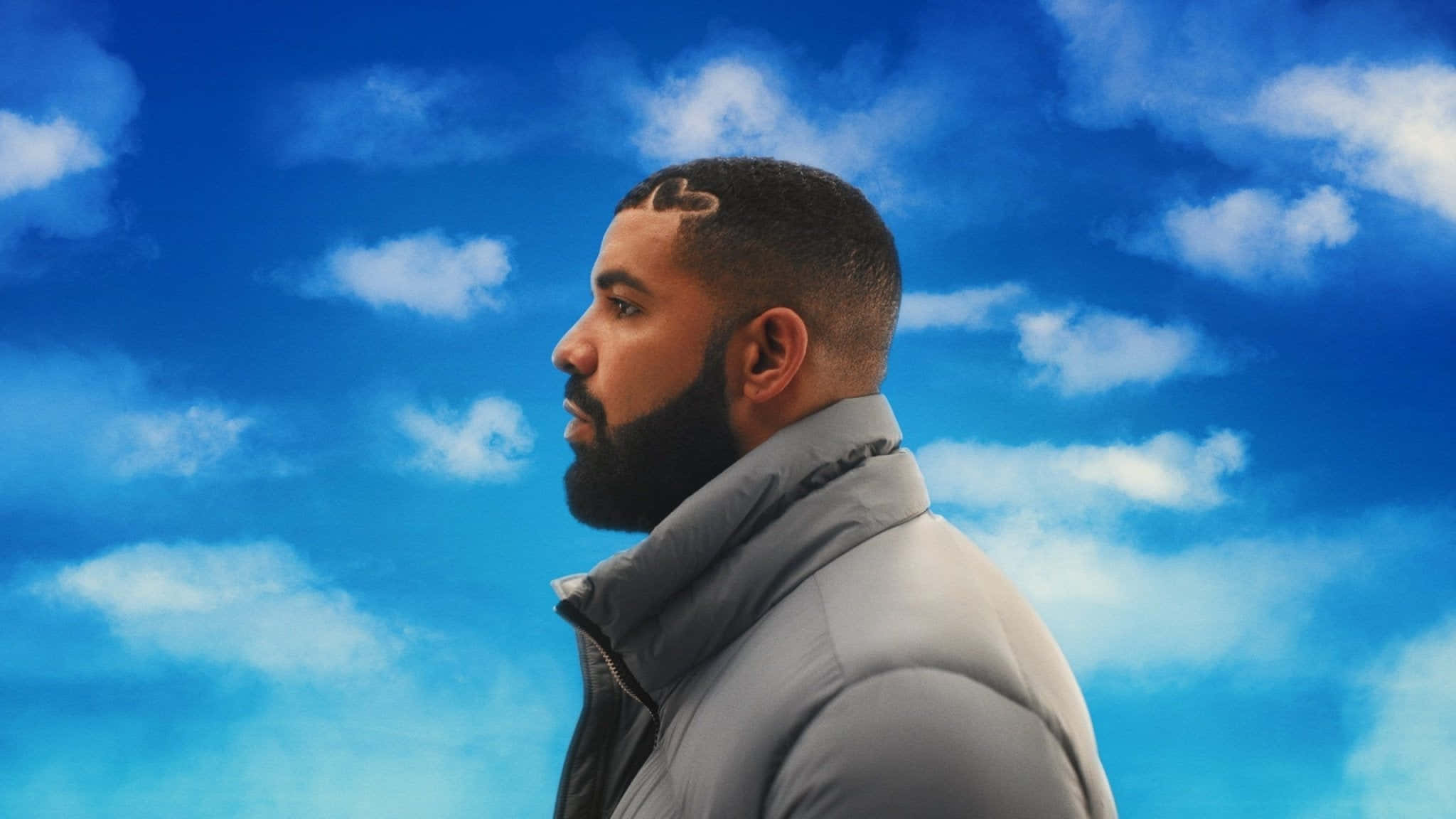 Drake - 《Nothing Was The Same》 壁紙 — 一個留鬍鬚、身穿灰色外套的男人，在藍天白雲的背景下側面站立。