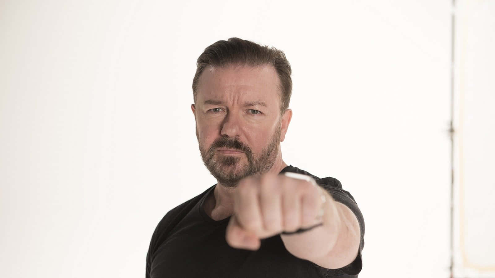 ricky gervais 桌布 壁紙 — 一個留鬍鬚、身穿黑色恤衫的男人，在純白背景前直接指向觀眾。