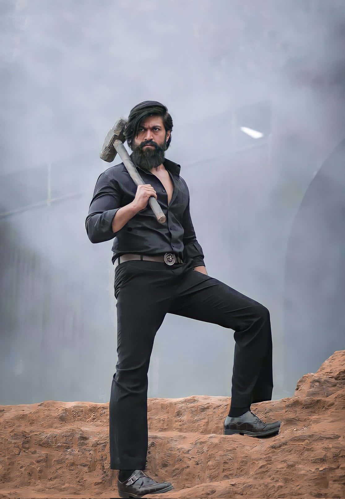 Kgf 2 桌布 壁紙 — 一個留鬍鬚、身穿黑色服裝的男人，在煙霧瀰漫的環境中，強而有力地站在岩石地形上，手持一把大錘。