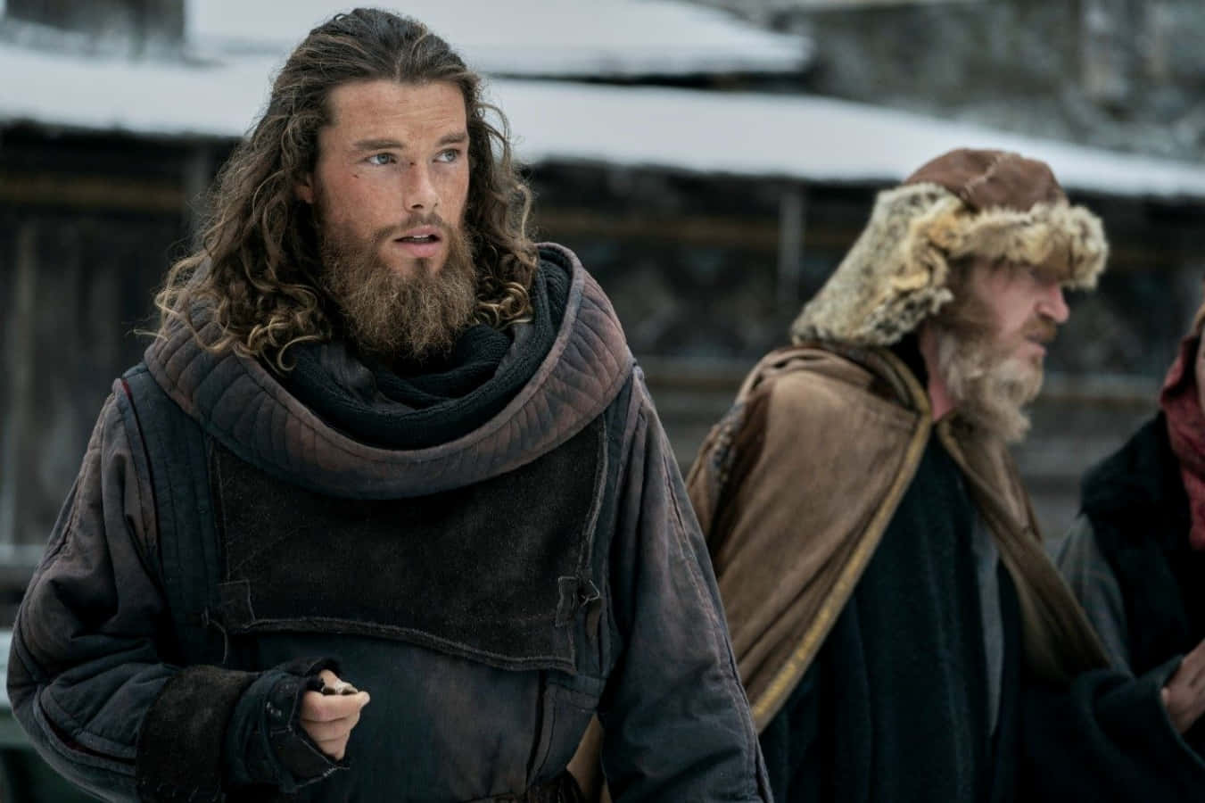 Vikings 桌布 壁紙 — 一個留鬍鬚嘅男人穿着粗獷嘅冬天衫，喺一個落雪嘅村莊警覺咁企喺度，旁邊仲有另一個人。