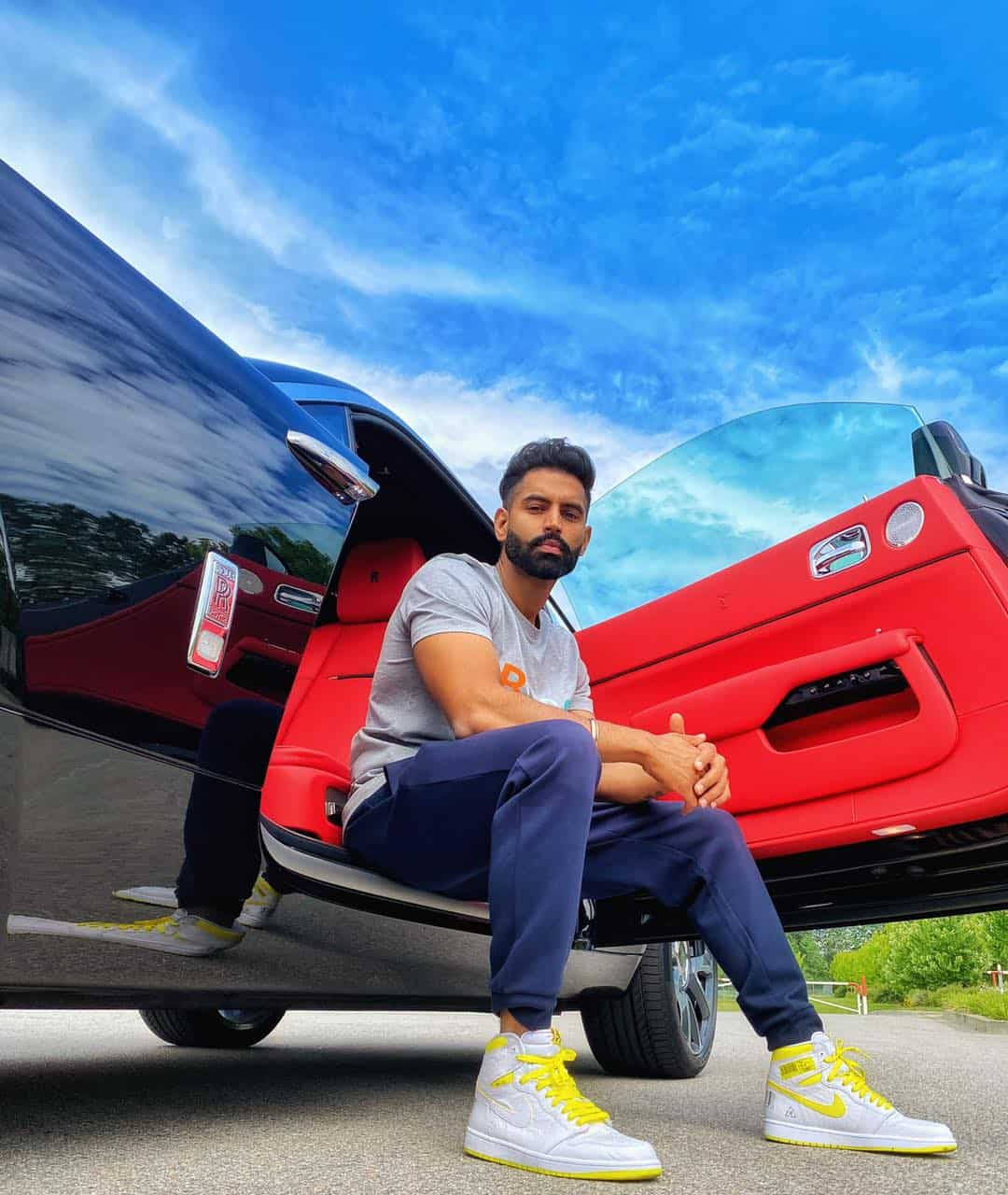 Indian 桌布 壁紙 — 一個留鬍鬚嘅男人自信咁坐喺一架紅色同黑色跑車嘅打開門上面，背景係晴朗藍天同散雲。
