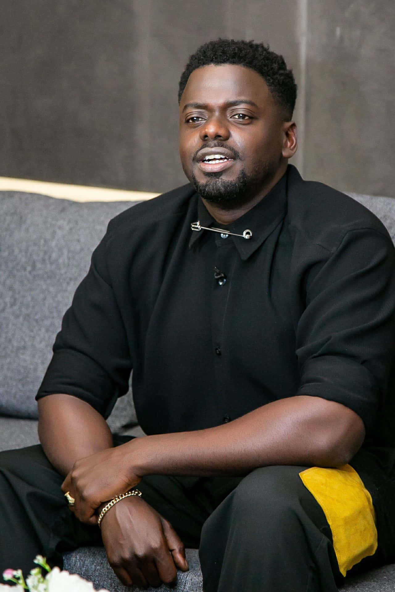Daniel Kaluuya 桌布 壁紙 — 一個留鬍鬚的男人放鬆地坐在灰色梳化上，穿著黑色襯衫，配有銀色別針和金色飾物。
