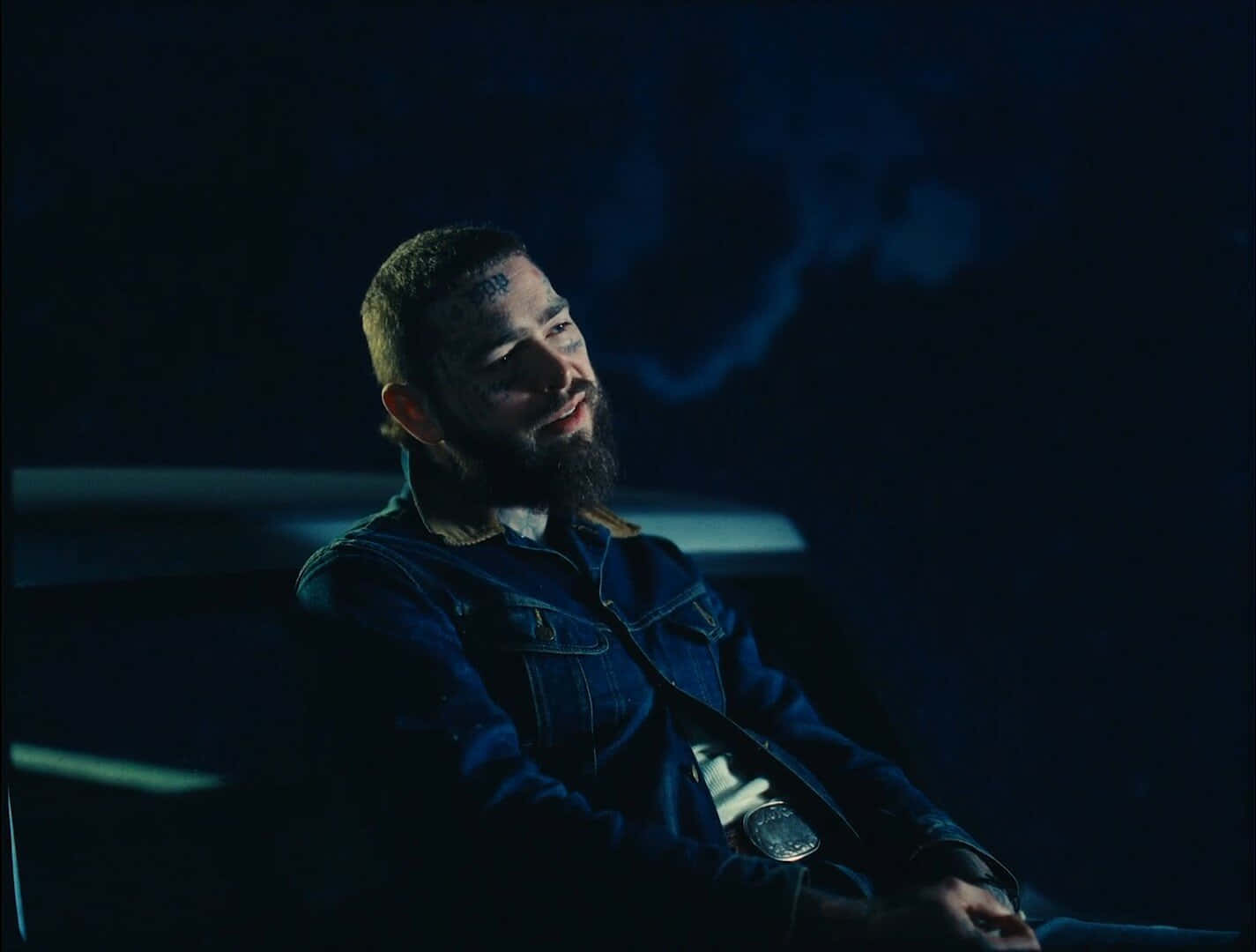Post Malone 桌布 壁紙 — 一個留鬍鬚的男人穿著牛仔外套坐在黑暗的車內，被冷藍色的光照亮，身後煙霧繚繞。