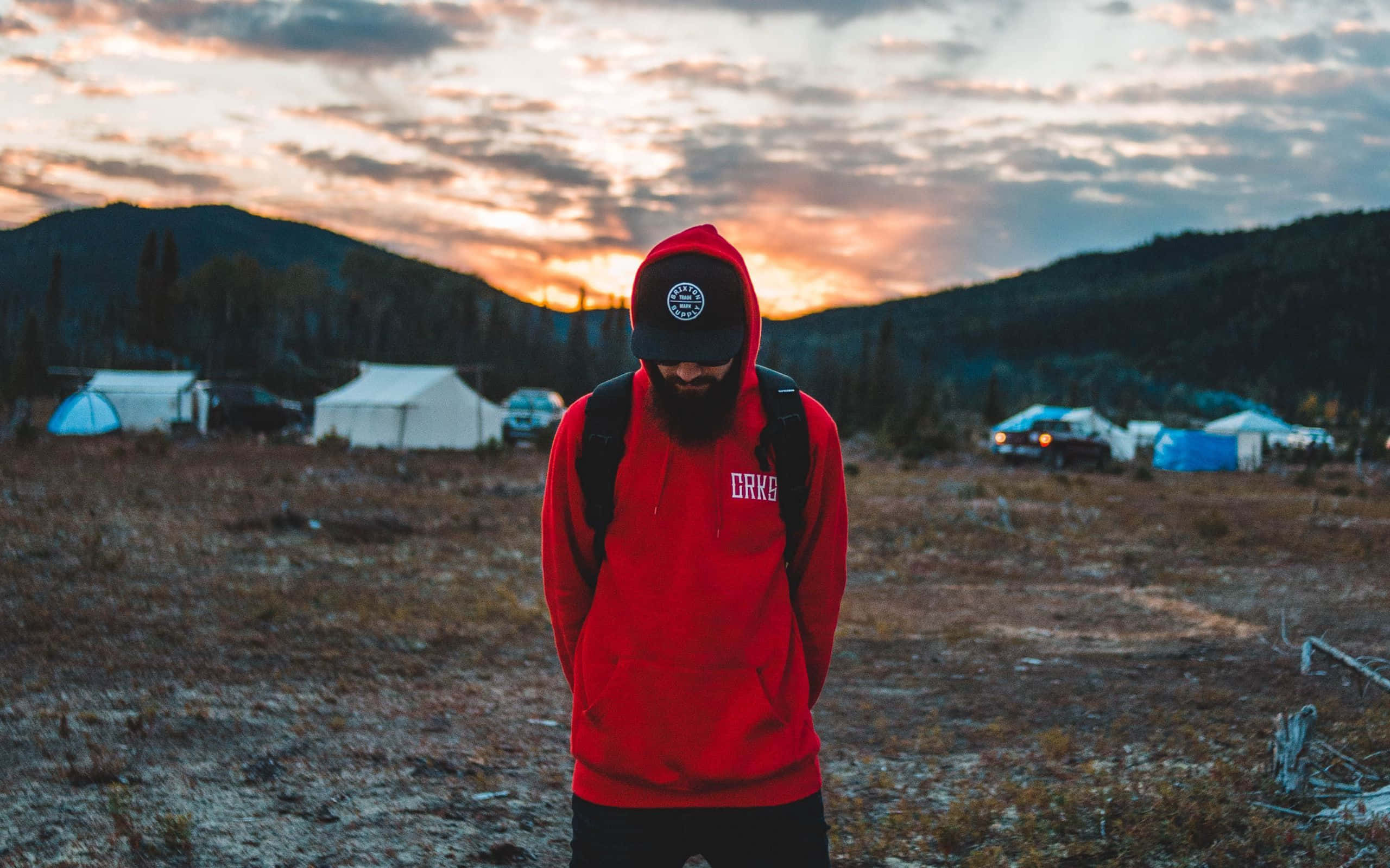 Red Hoodie 桌布 壁紙 — 一個留鬍鬚的男人穿著紅色連帽衫，在日落時分站在田野裡，背景有帳篷和山脈。