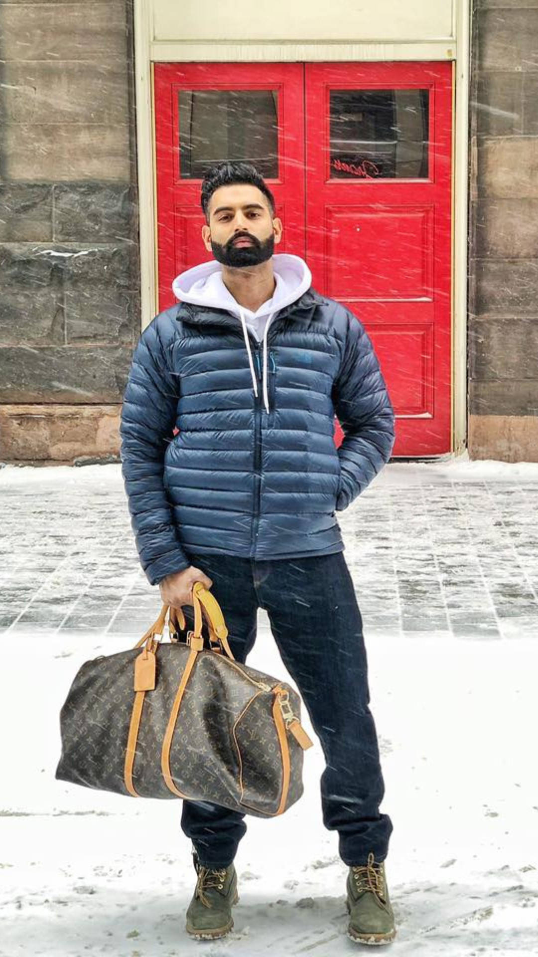Indian 桌布 壁紙 — 一個留鬍鬚的男人穿著藍色羽絨外套，站在飄雪中，在紅色門前拿著一個路易威登行李袋。