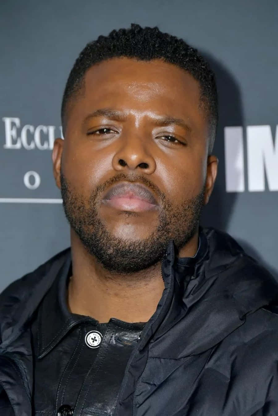 Winston Duke 桌布 壁紙 — 一個留鬍鬚的男人穿著黑色外套，以深灰色背景和部分白色字體的特寫肖像。