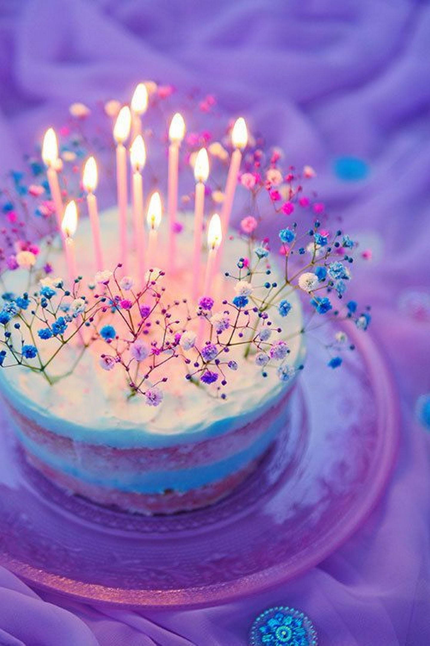 生日蛋糕 桌布 壁紙 — 一個異想天開的生日蛋糕，帶有發光的蠟燭和柔和的滿天星花朵，放在紫色布料背景上。