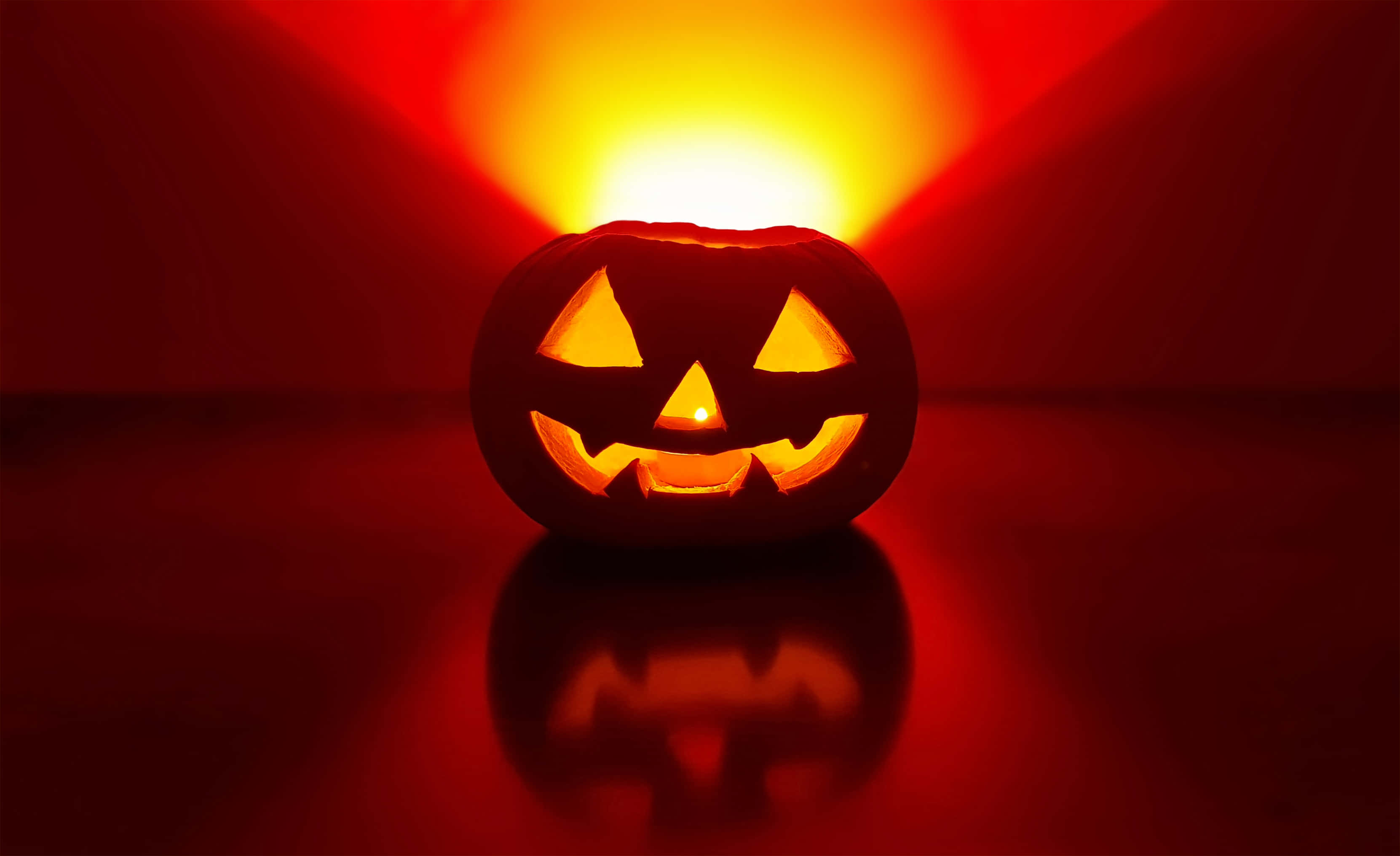Halloween Crafts 桌布 壁紙 — 一個發光的傑克南瓜燈，裏面有蠟燭，在帶有反光表面的深紅色背景上投射出溫暖的橙色光芒。