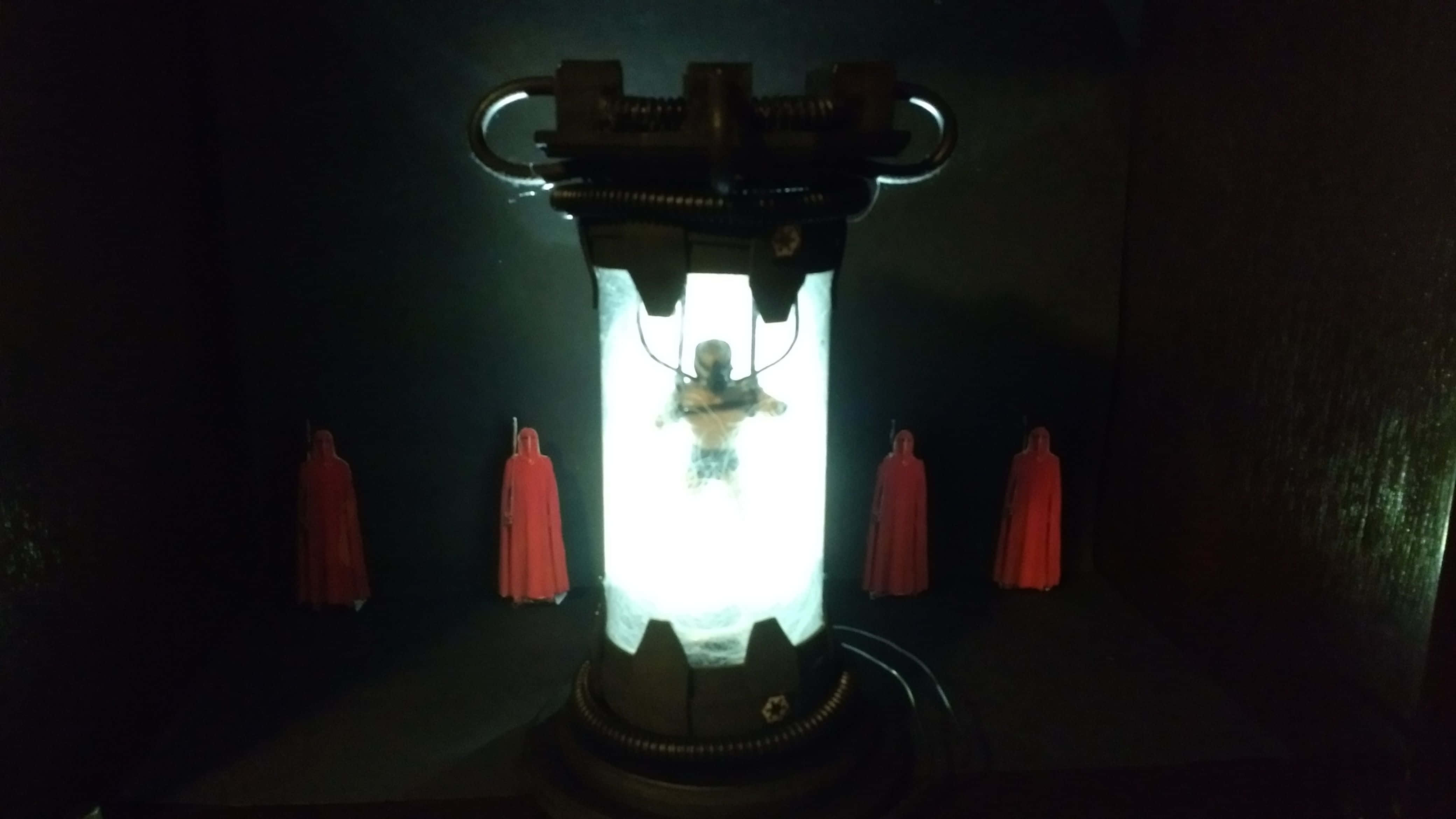 Bacta Tank 桌布 壁紙 — 一個發光的圓柱形水箱內懸浮著一個類人形體，四個身穿紅袍的人影站在周圍的黑暗中。