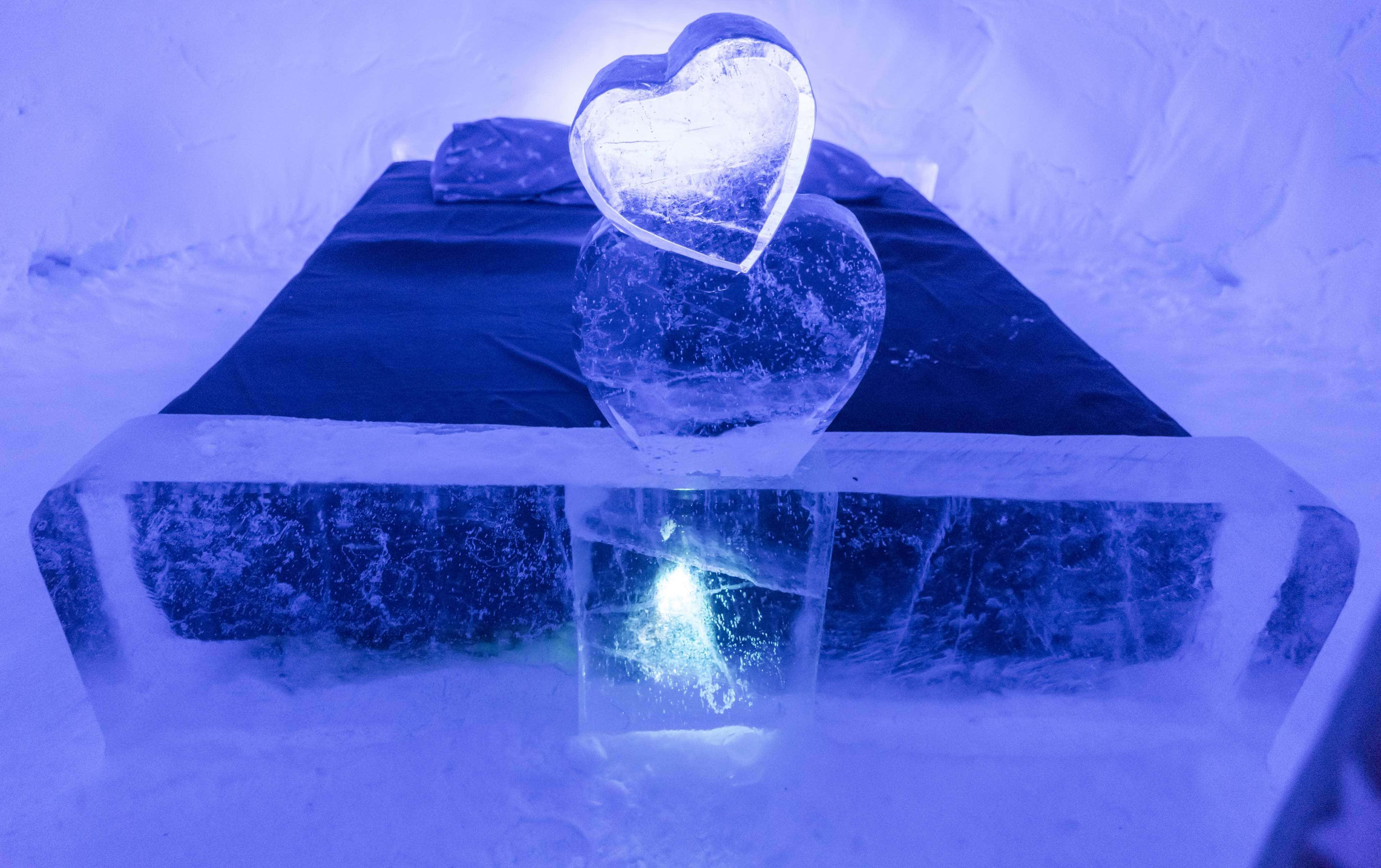 Ice Sculpture 桌布 壁紙 — 一個發光的愛心形狀冰雕放在一張床上，房間是藍色燈光的冰室，牆壁覆蓋著雪。