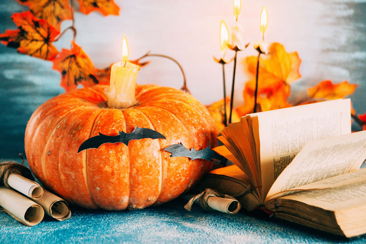 Halloween Books 桌布 壁紙 — 一個發光的橙色南瓜，帶有蝙蝠鏤空和蠟燭，旁邊放著一本打開的書和秋葉，在帶紋理的藍色表面上。