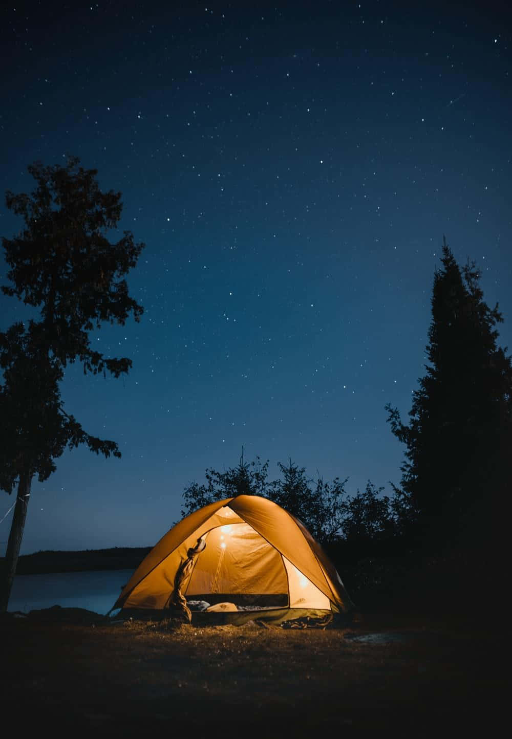 Beach Camping 桌布 壁紙 — 一個發光的橙色帳篷，坐落在湖邊，上方是繁星點點的夜空，四周環繞著深色樹木。