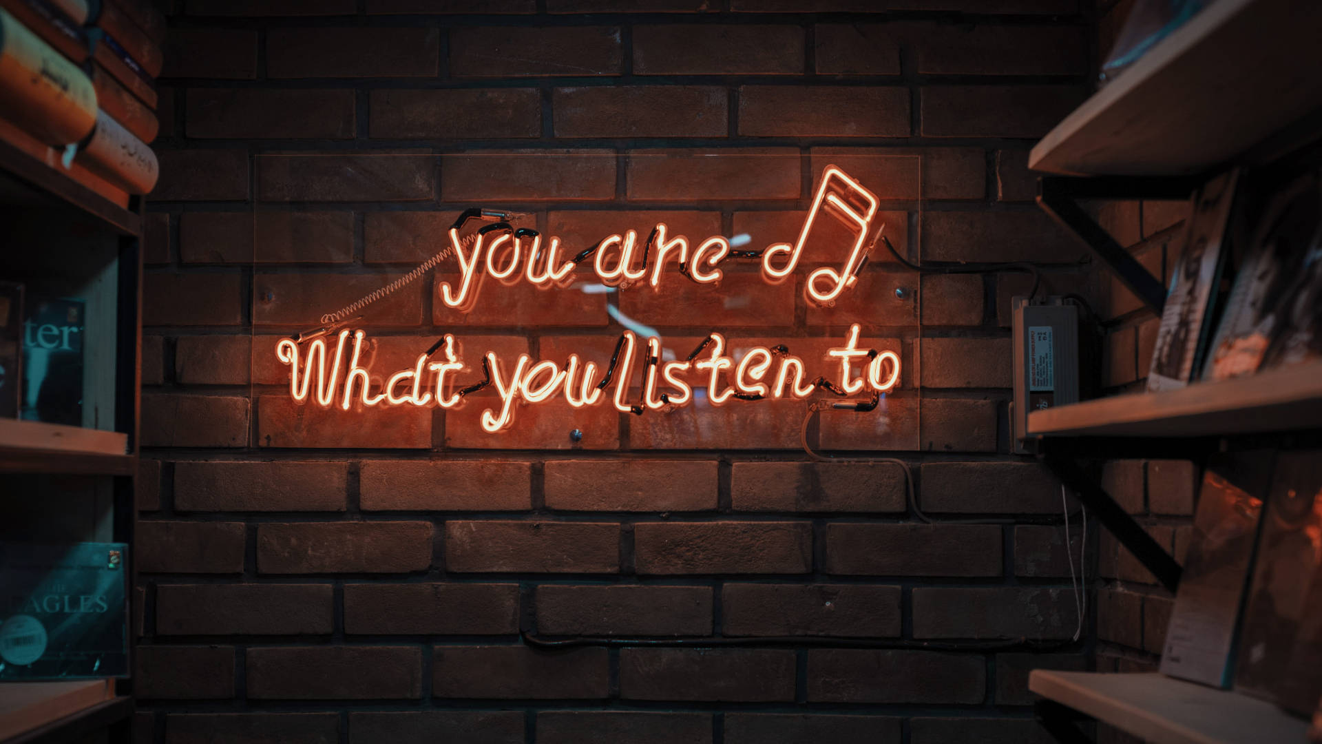 Brick 桌布 壁紙 — 一個發光的橙色霓虹燈牌，上面寫著「you are what you listen to」並有一個音符，掛在深色磚牆上，兩旁是木製架子。