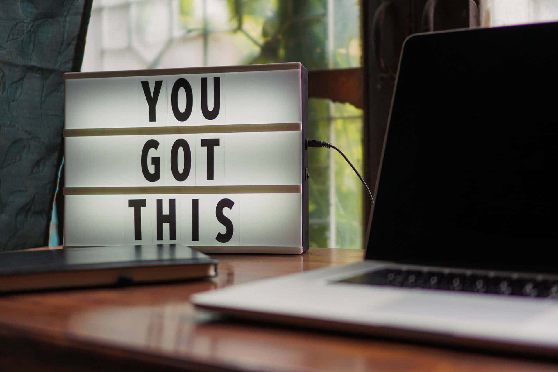 Positive 桌布 壁紙 — 一個發光的燈箱顯示著「YOU GOT THIS」，放在靠近窗戶、有綠色植物的木桌上，旁邊有一部手提電腦。