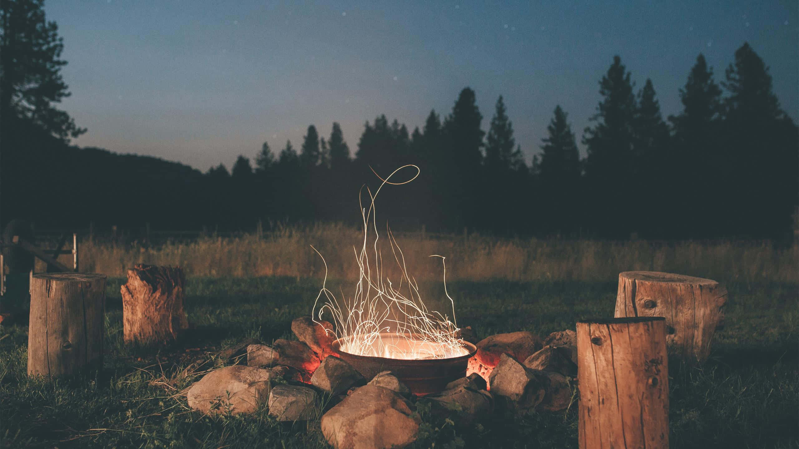 Cozy Fire 桌布 壁紙 — 一個發光的營火，火花舞動，坐在一個由樹樁包圍的石環中，背景是星光閃爍的黃昏天空。