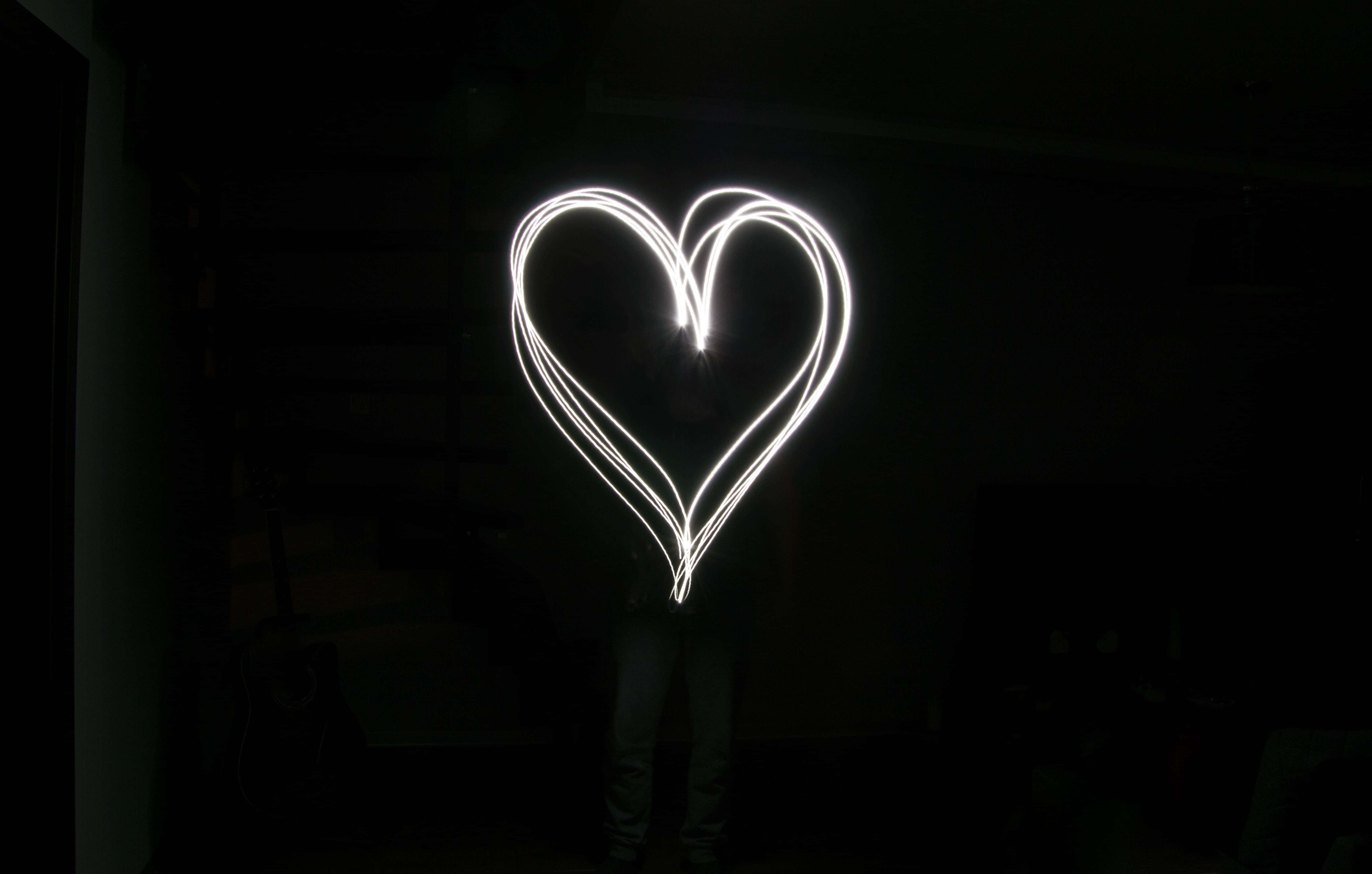 Black And White Heart 桌布 壁紙 — 一個發光的白色心形，由一個人站在昏暗的房間中，用光線繪畫而成，房間內有模糊的家具輪廓。