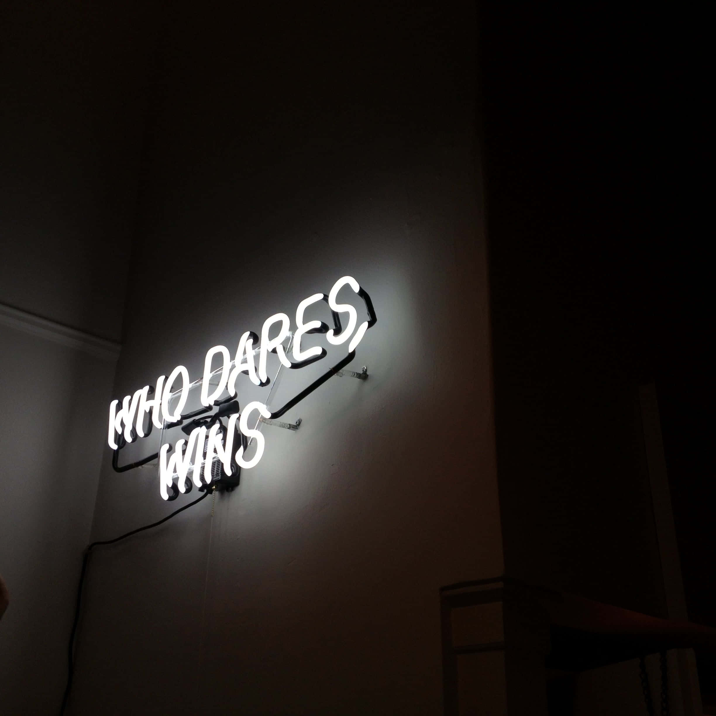 Daring 桌布 壁紙 — 一個發光的白色霓虹燈牌，上面寫著「WHO DARES WINS」，照亮了陰暗城市室內環境中的一面深色牆壁。