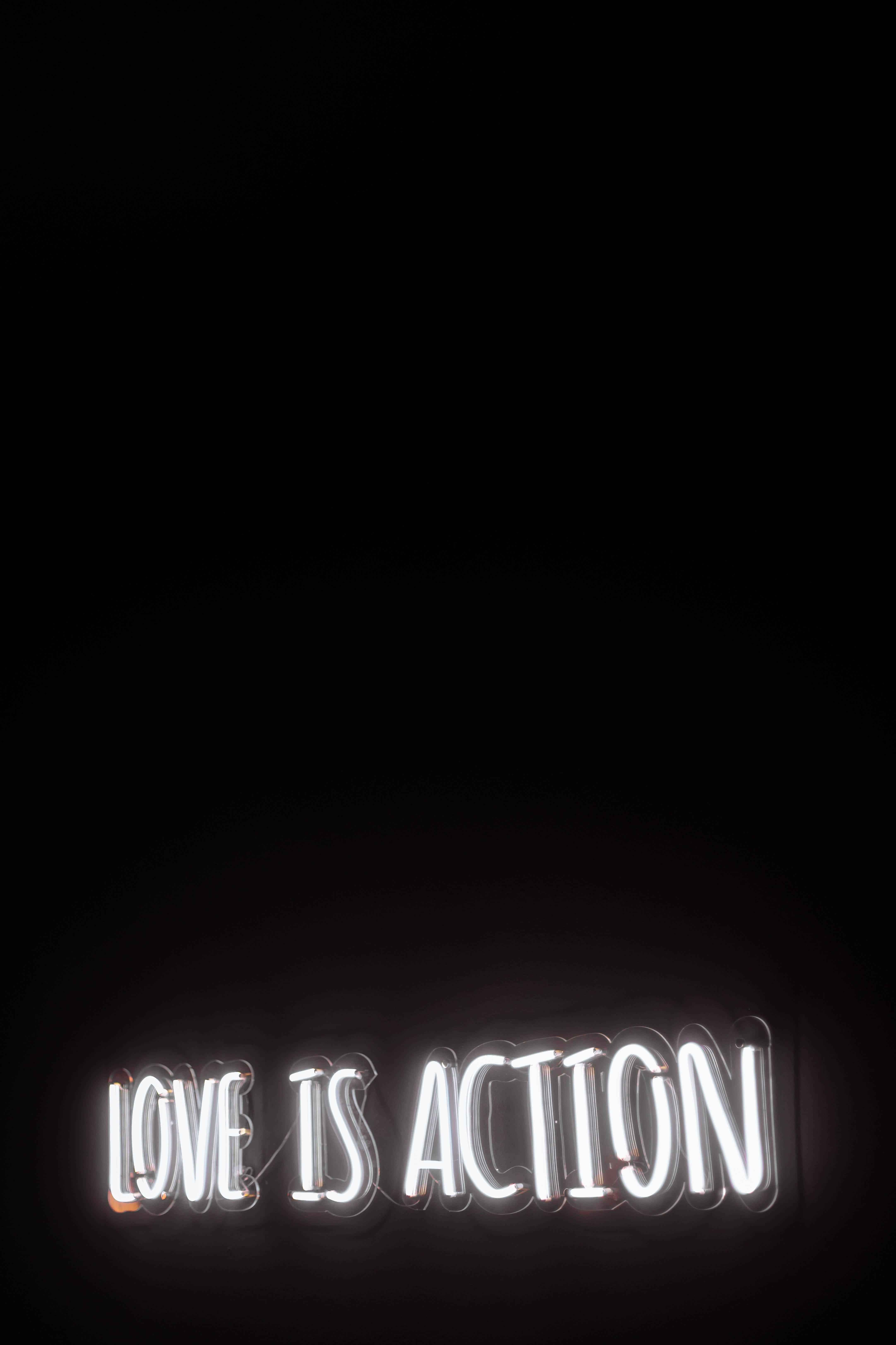 愛心 桌布 壁紙 — 一個發光的白色霓虹燈牌，上面寫著 love is action，背景是漆黑的。