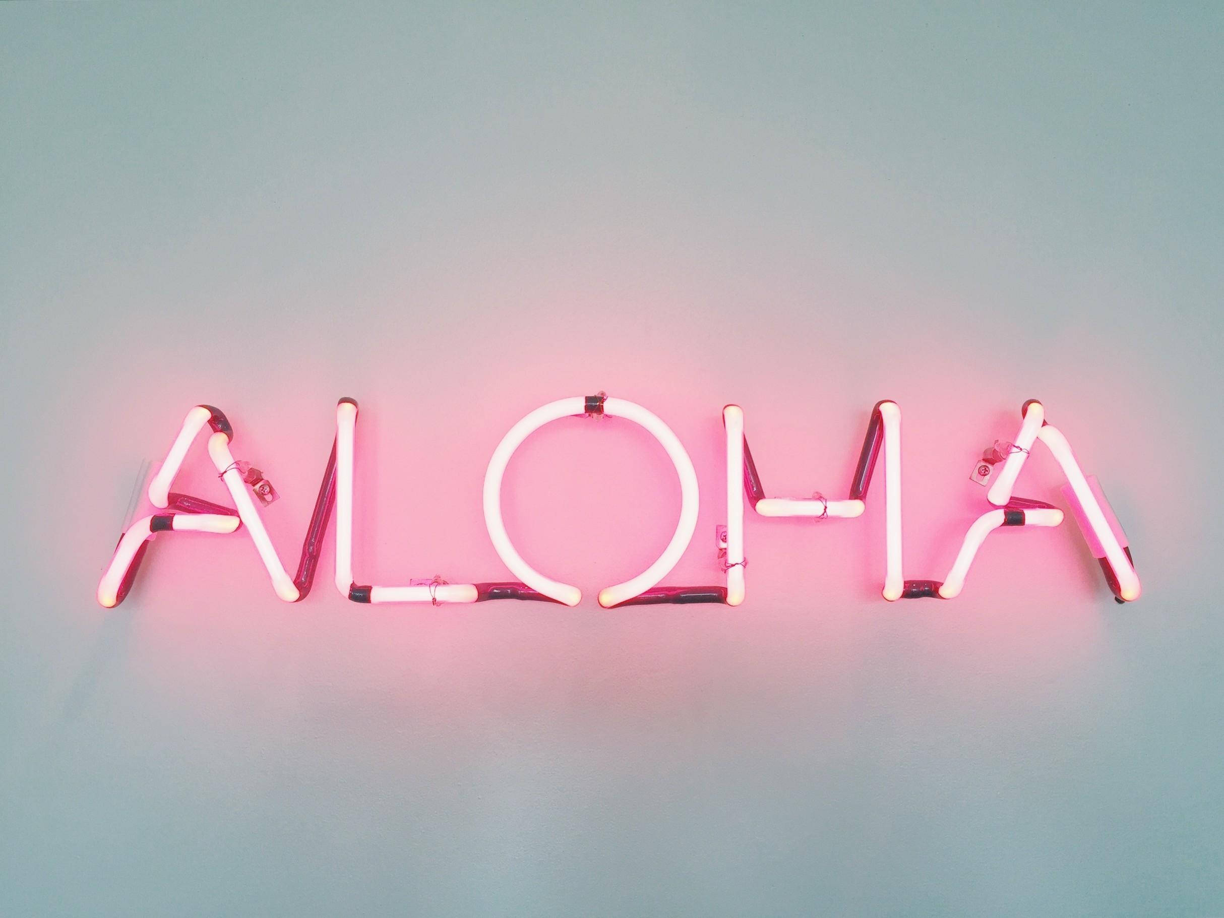 indie aesthetic laptop 桌布 壁紙 — 一個發光的粉紅色霓虹燈牌，上面拼寫著「ALOHA」，在柔和的青綠色牆壁上投射出柔和的光芒。