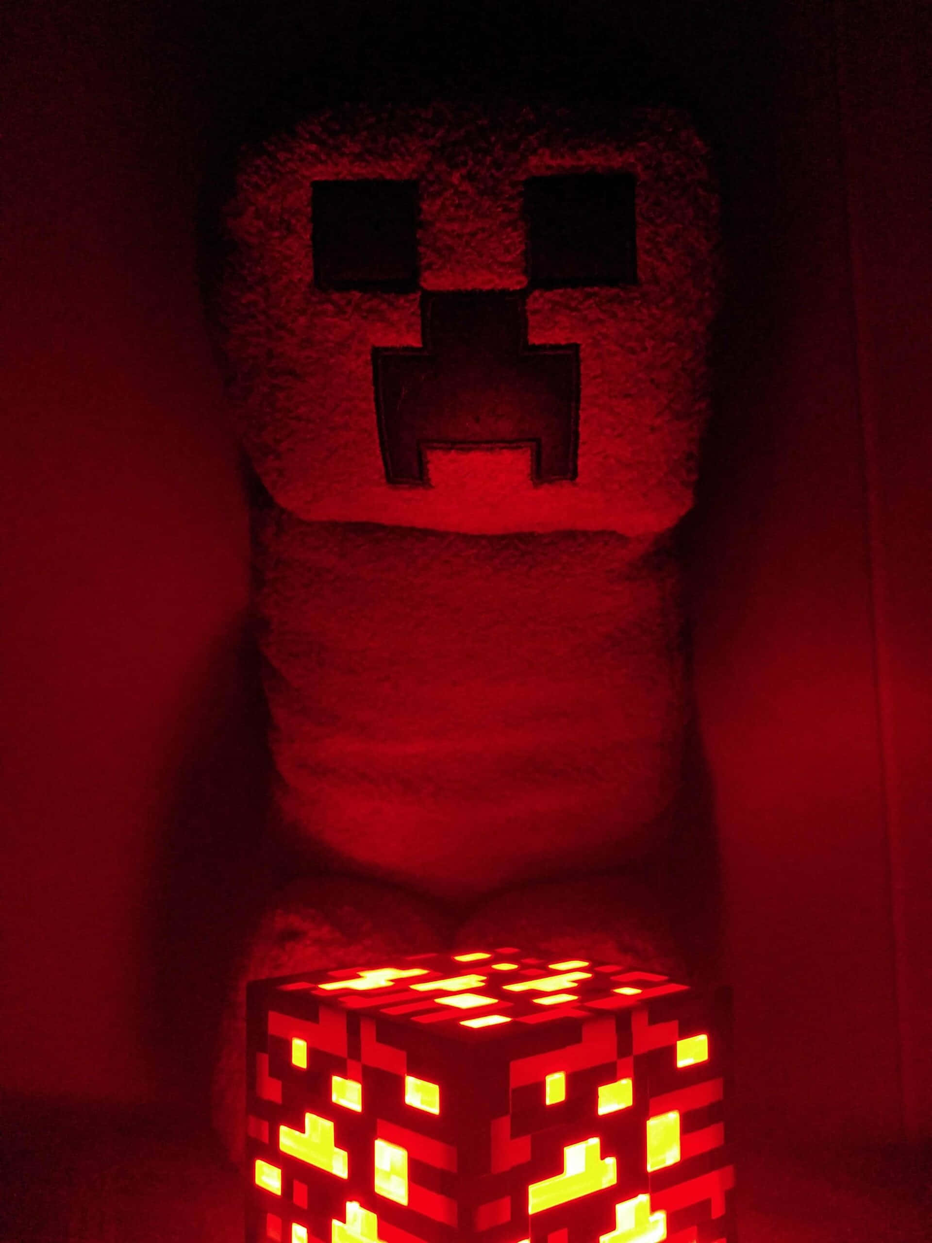 Minecraft Redstone 桌布 壁紙 — 一個發光的紅色Minecraft Creeper毛絨玩具站在一個像素化的Creeper燈後面，在黑暗的房間中投射出溫暖的光線。