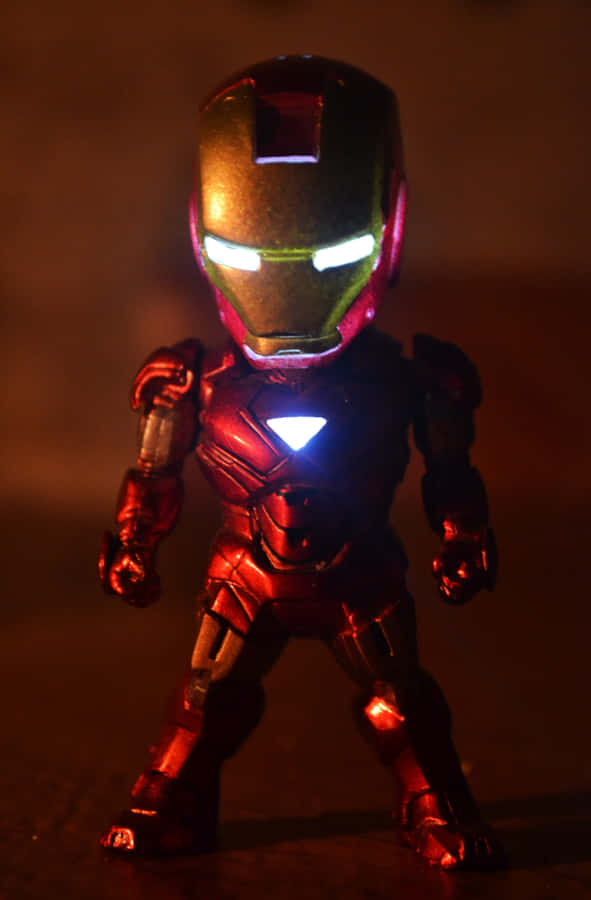 Iron Man Action Figures 桌布 壁紙 — 一個發光的紅色和金色鐵甲奇俠動作人偶站在昏暗的燈光下，眼睛和胸部弧形反應堆發光。