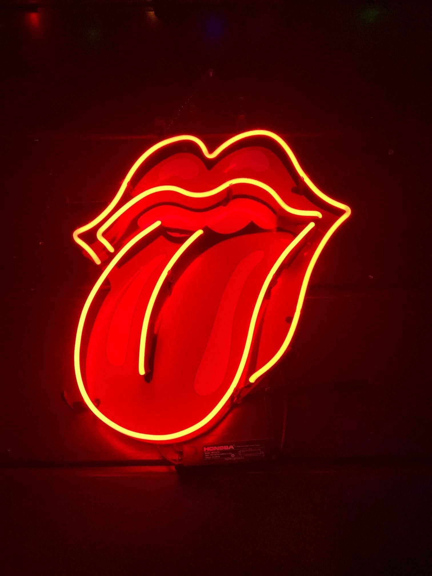 Tongue Out 桌布 壁紙 — 一個發光的紅色霓虹燈標誌，顯示舌頭從嘴唇伸出，以鮮豔的橙色輪廓照亮黑暗的牆壁。