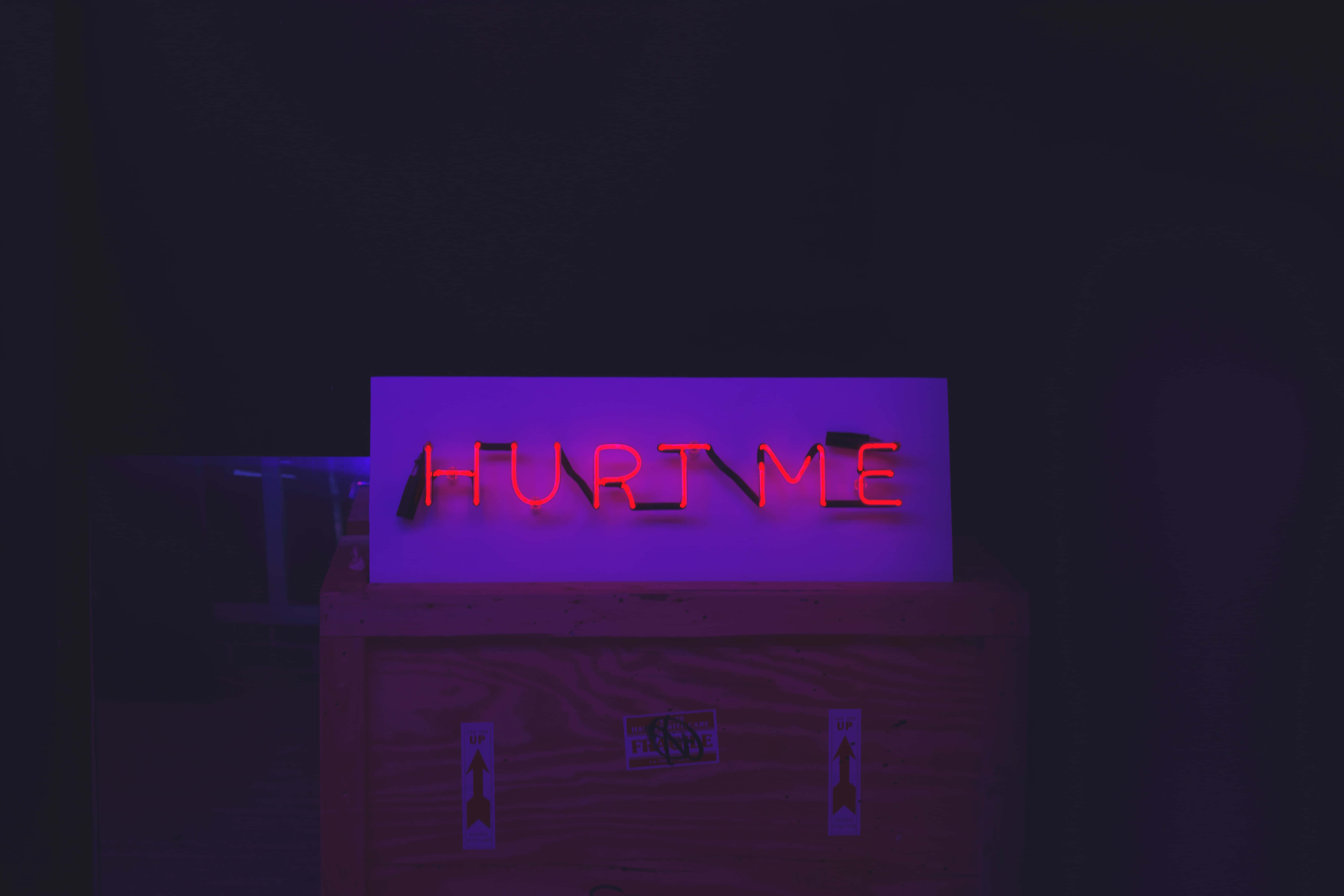 hurt 桌布 壁紙 — 一個發光的紅色霓虹燈牌，上面寫著「HURT ME」，在黑暗的房間中將紫色色調投射在木箱上。
