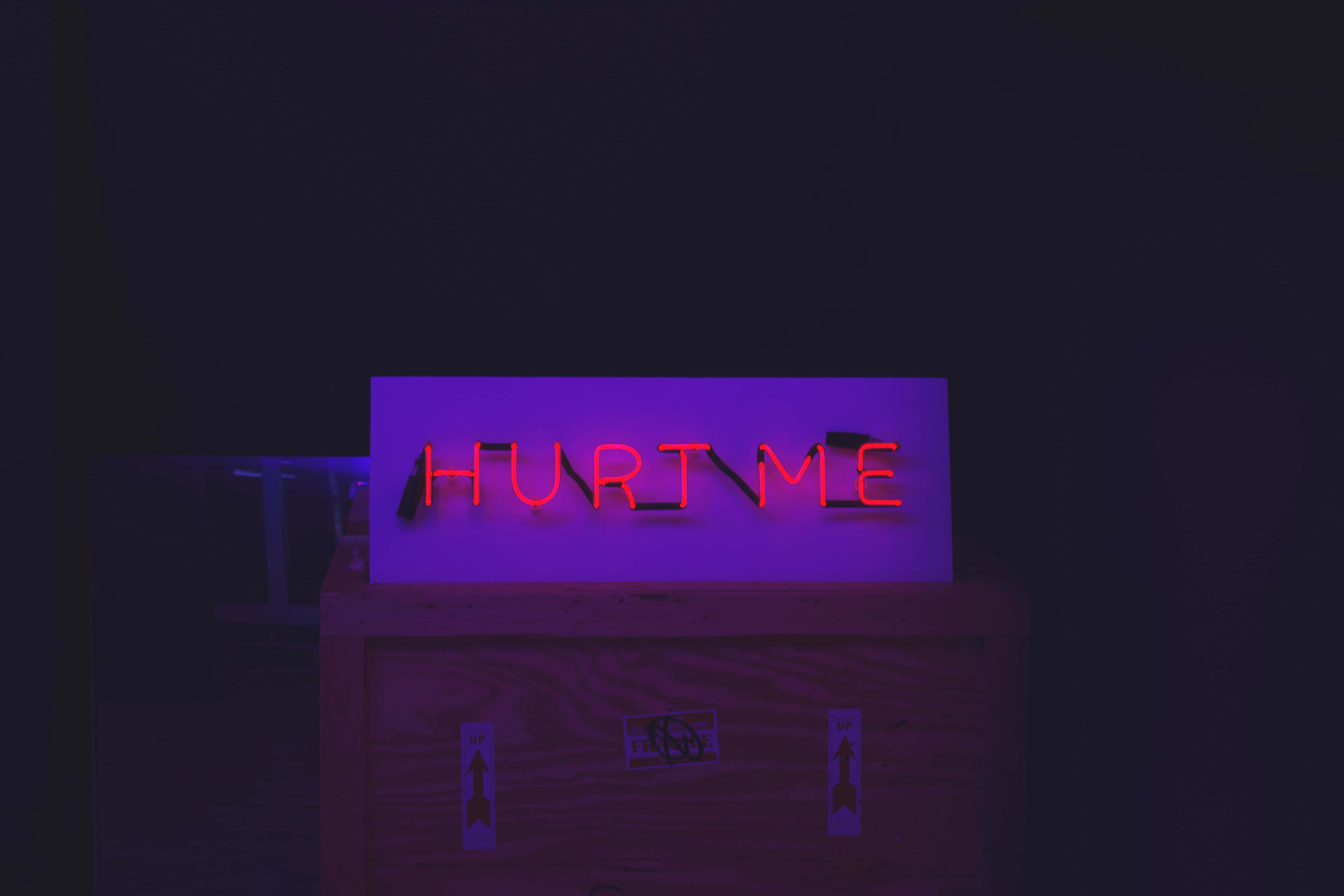 neon sign 桌布 壁紙 — 一個發光的紅色霓虹燈牌，上面寫著「HURT ME」，在黑暗的房間中將紫色光暈投射在木箱上。