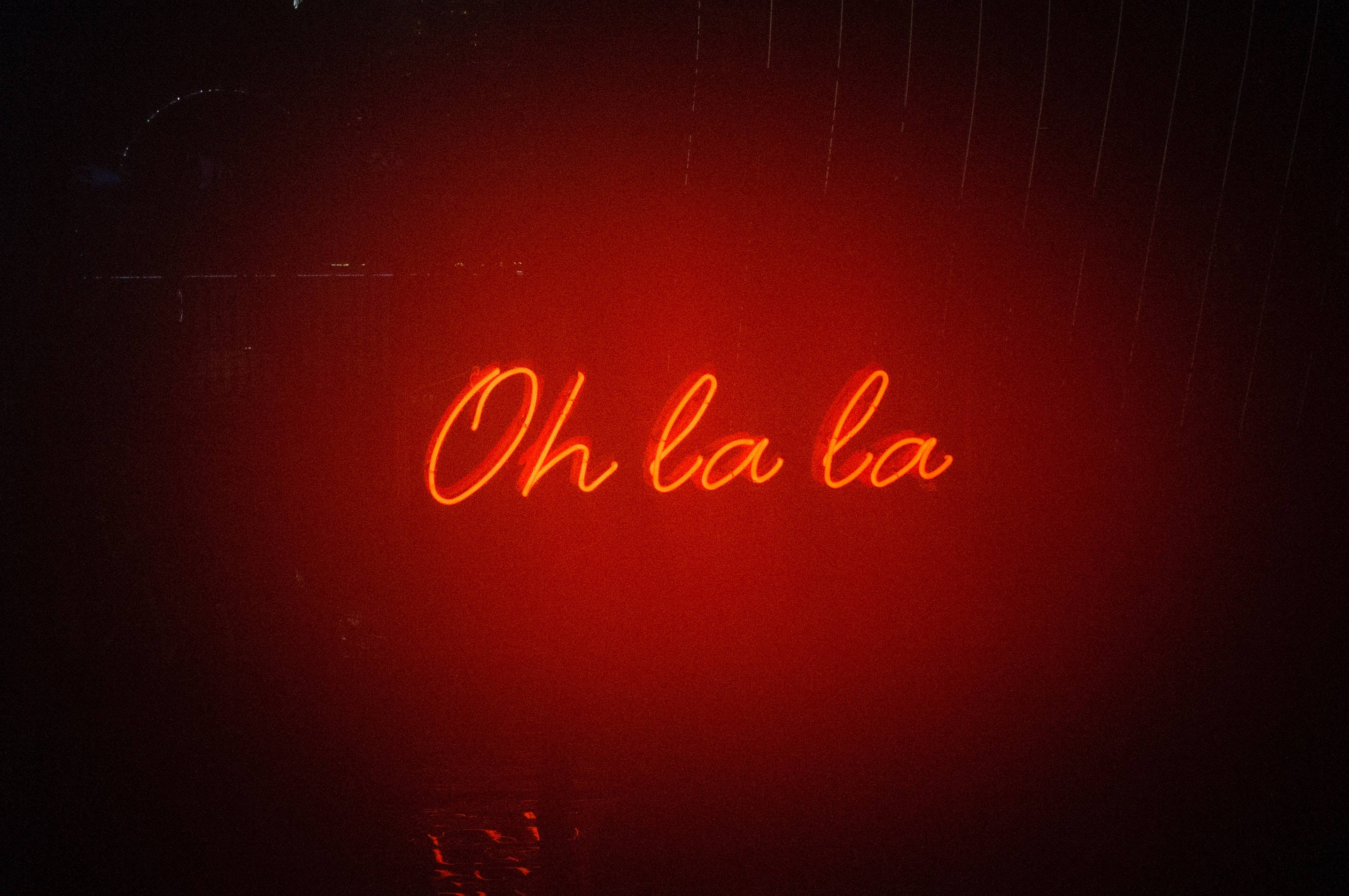 黑紅 桌布 壁紙 — 一個發光的紅色霓虹燈牌，上面寫著 Oh la la，在黑暗的雨窗上投射出溫暖的光芒。