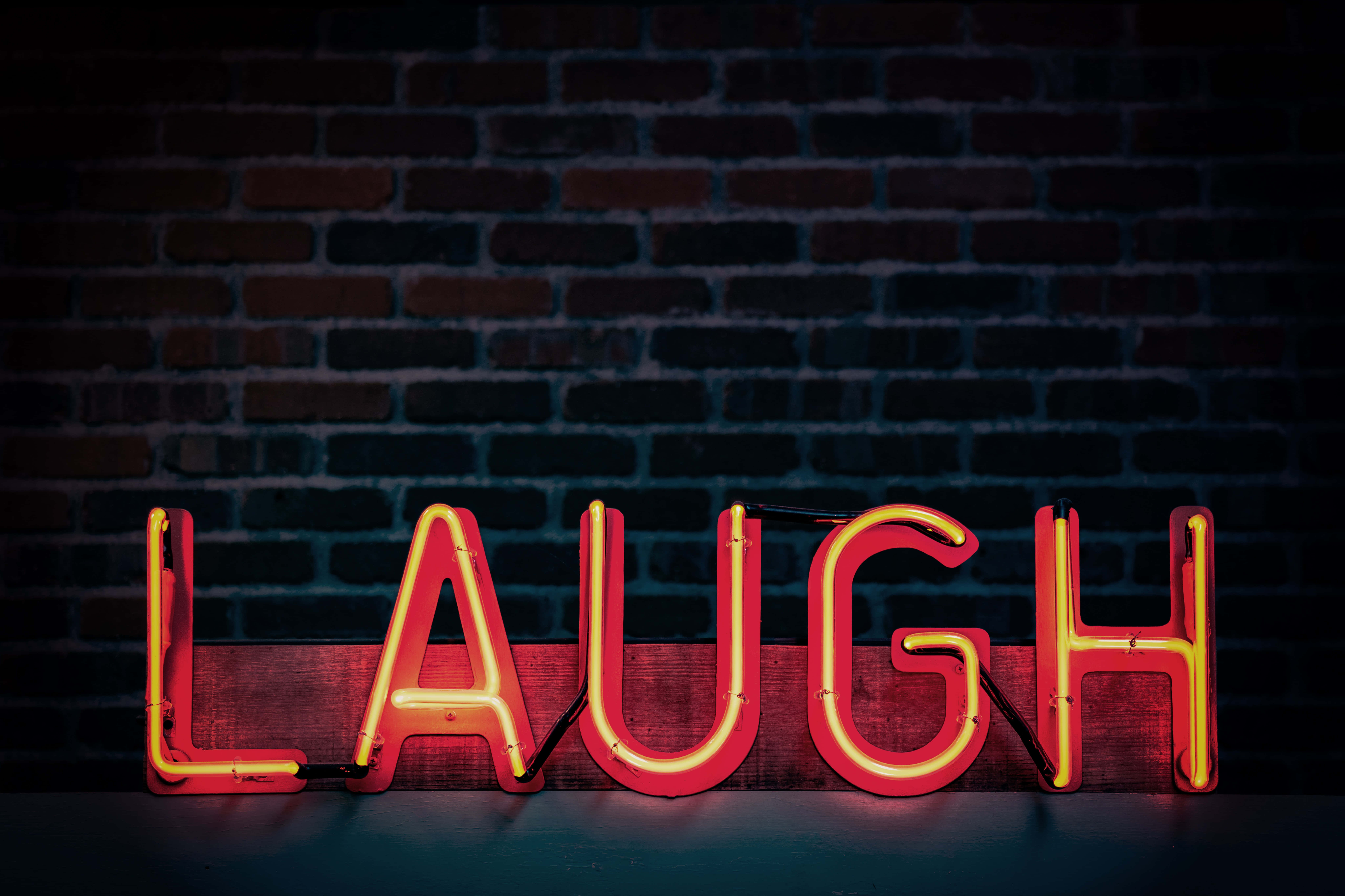 Neon Light 桌布 壁紙 — 一個發光的紅色霓虹燈牌，上面拼寫著 LAUGH，擱在木板上，背景是深色磚牆。