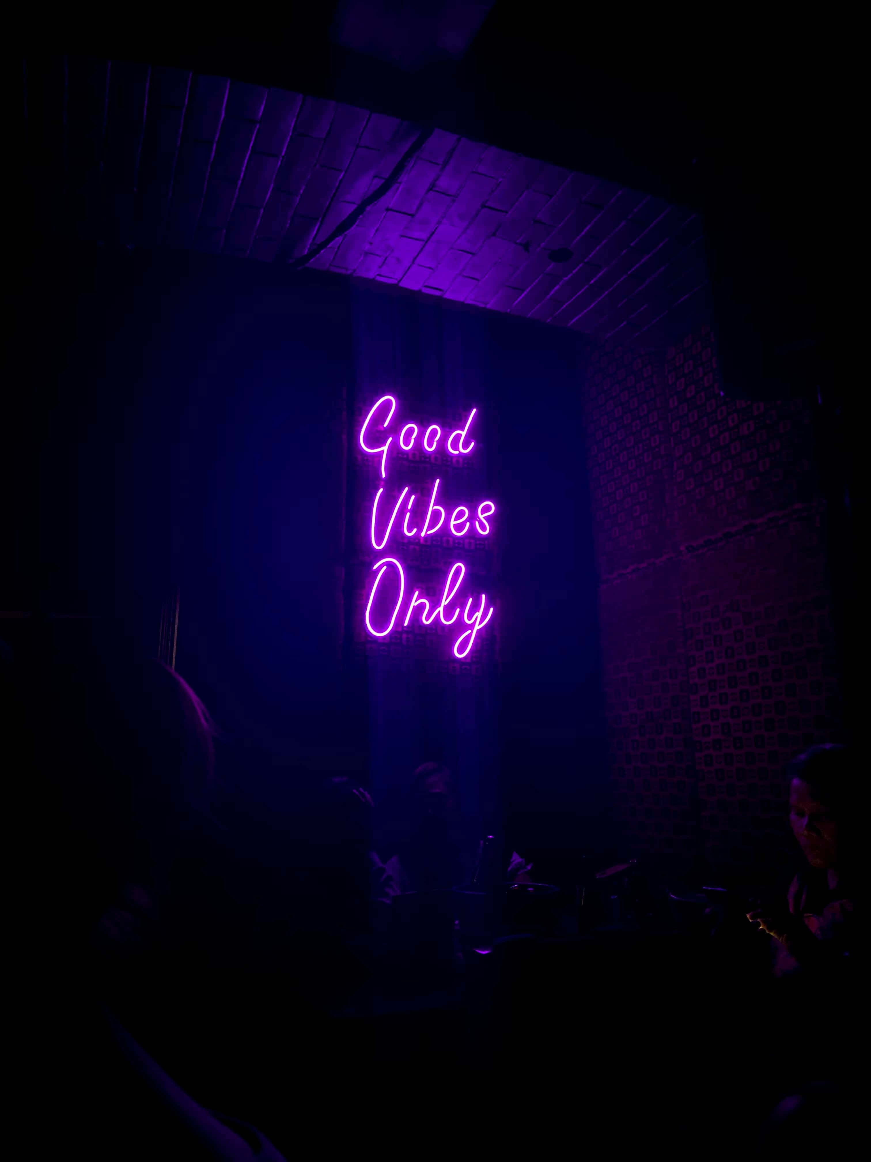 Vibes 桌布 壁紙 — 一個發光的紫色霓虹燈牌寫著 Good Vibes Only，照亮了一個光線昏暗、有磚牆和剪影顧客的房間。