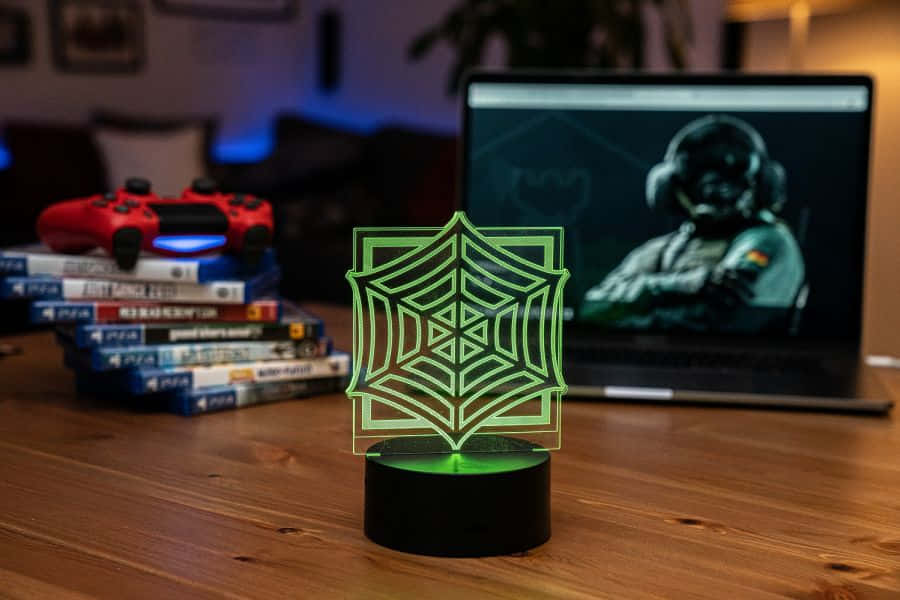 Rainbow Six Siege Jager 桌布 壁紙 — 一個發光的綠色蜘蛛網燈放在木桌上，旁邊有一個紅色PS5手掣和一部顯示遊戲玩家的手提電腦。